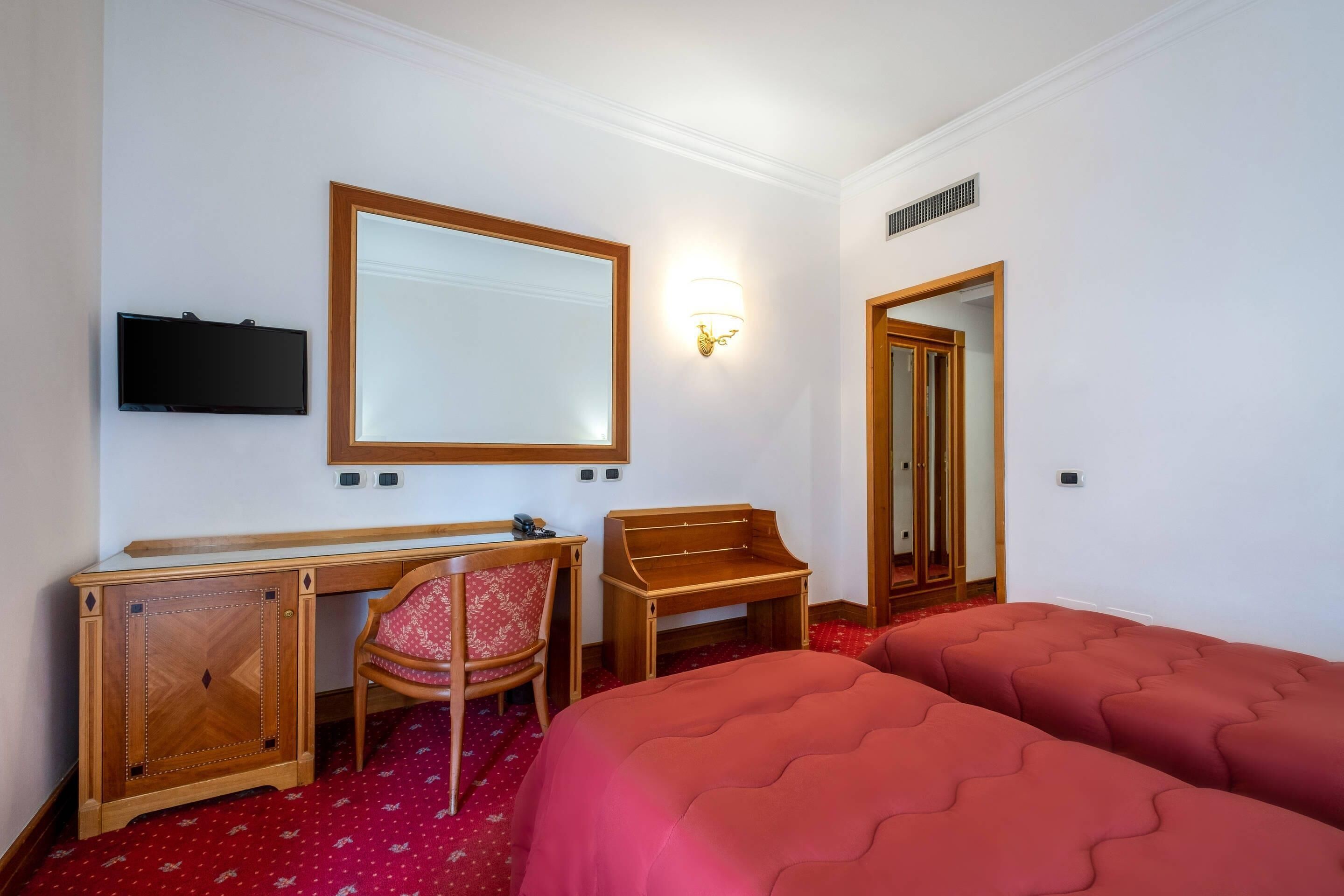 A hotel room with a desk, chair, two beds, a television, and a mirror.