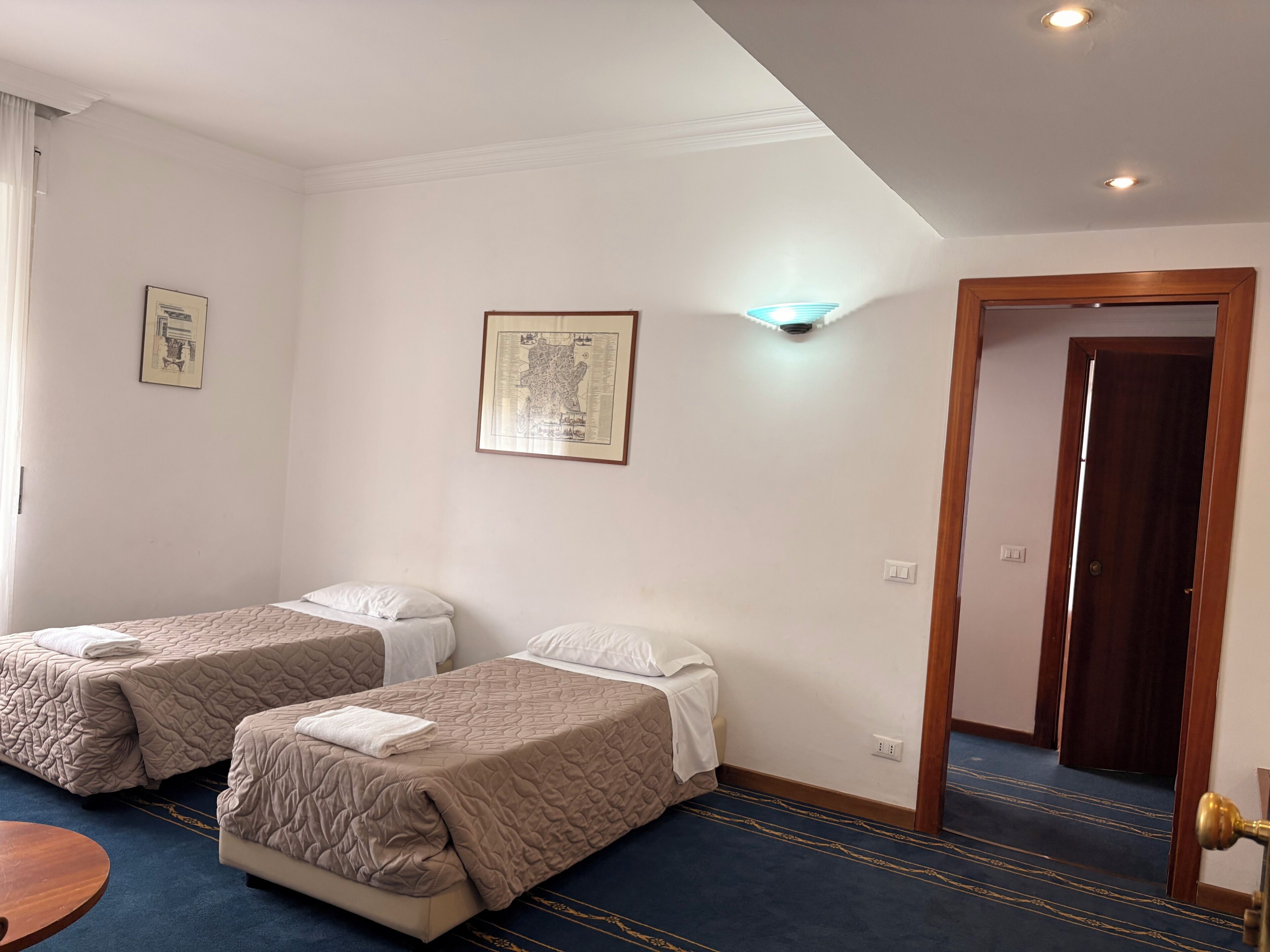 Habitación de hotel con dos camas, una mesita, un mapa enmarcado en la pared y una puerta que da a otra habitación.