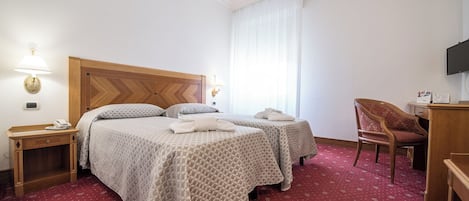A hotel room with a large bed, two bedside tables, a desk, a chair, a TV, and a window with curtains.