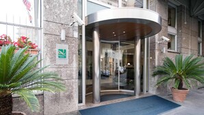 Entrada de hotel con puerta giratoria, felpudo azul y plantas en macetas.