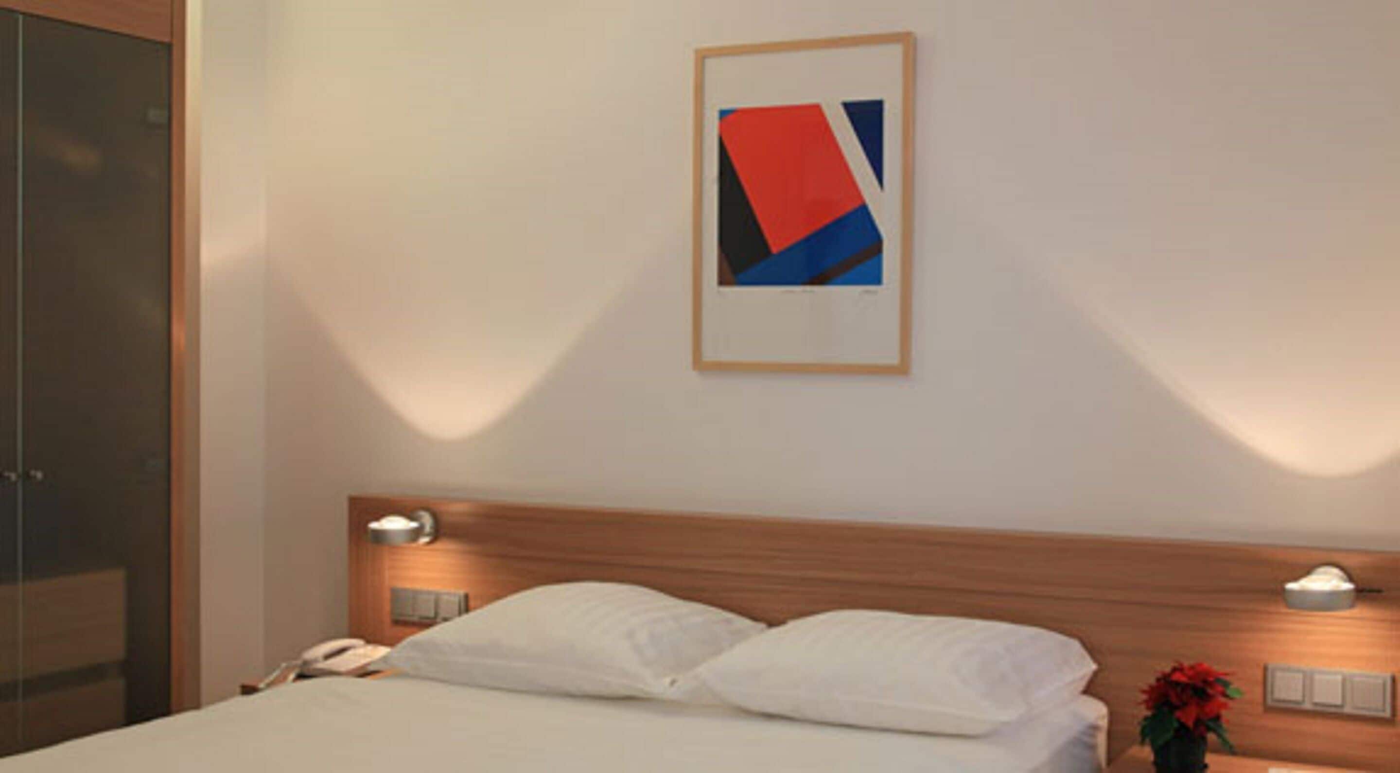 A hotel room with a bed, a framed abstract painting, bedside lamps, and a telephone on the nightstand.
