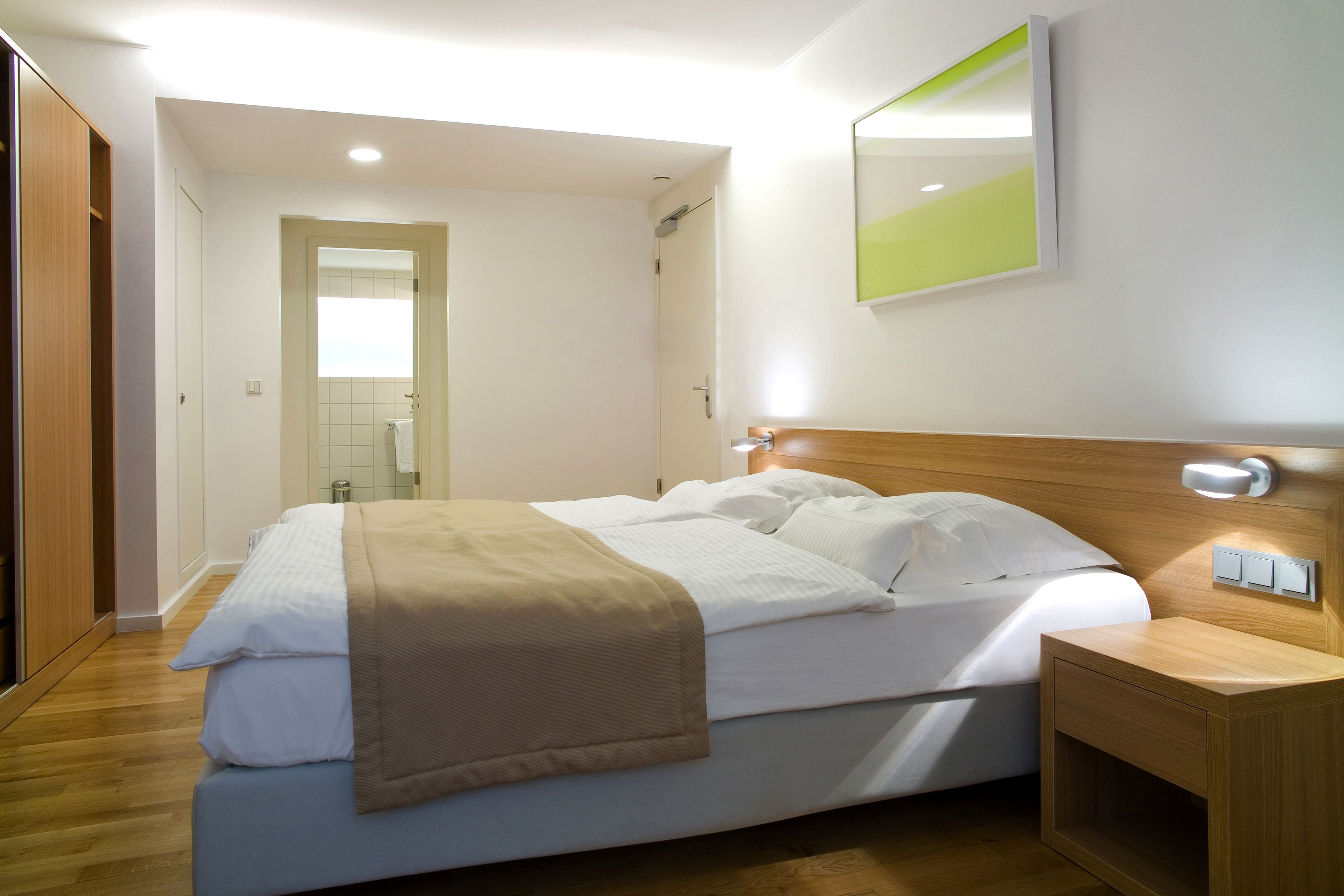 A modern hotel room with a large bed, wooden headboard, bedside table, and a mirror on the wall.
