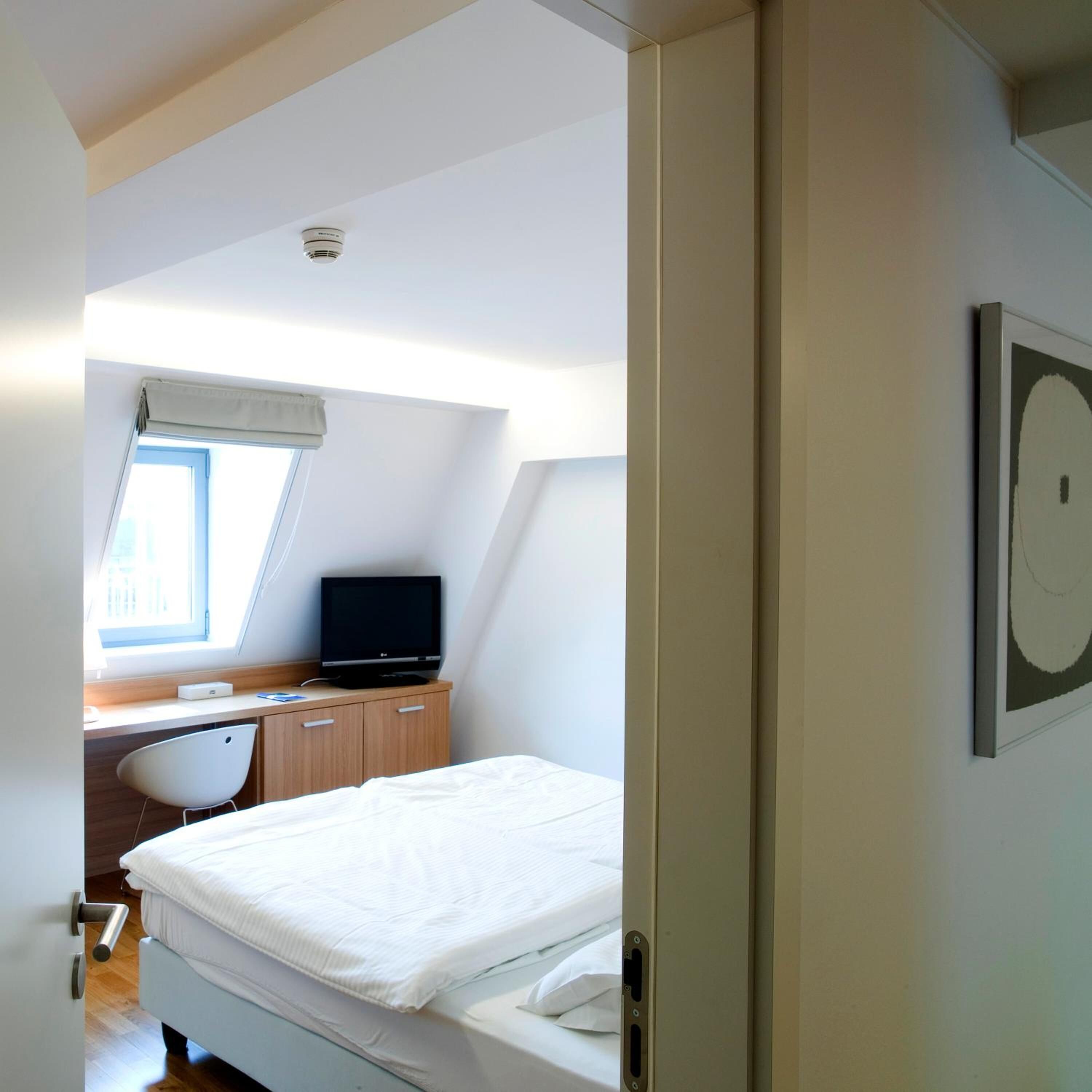 A bedroom with a bed, a desk with a computer, a chair, and a window.