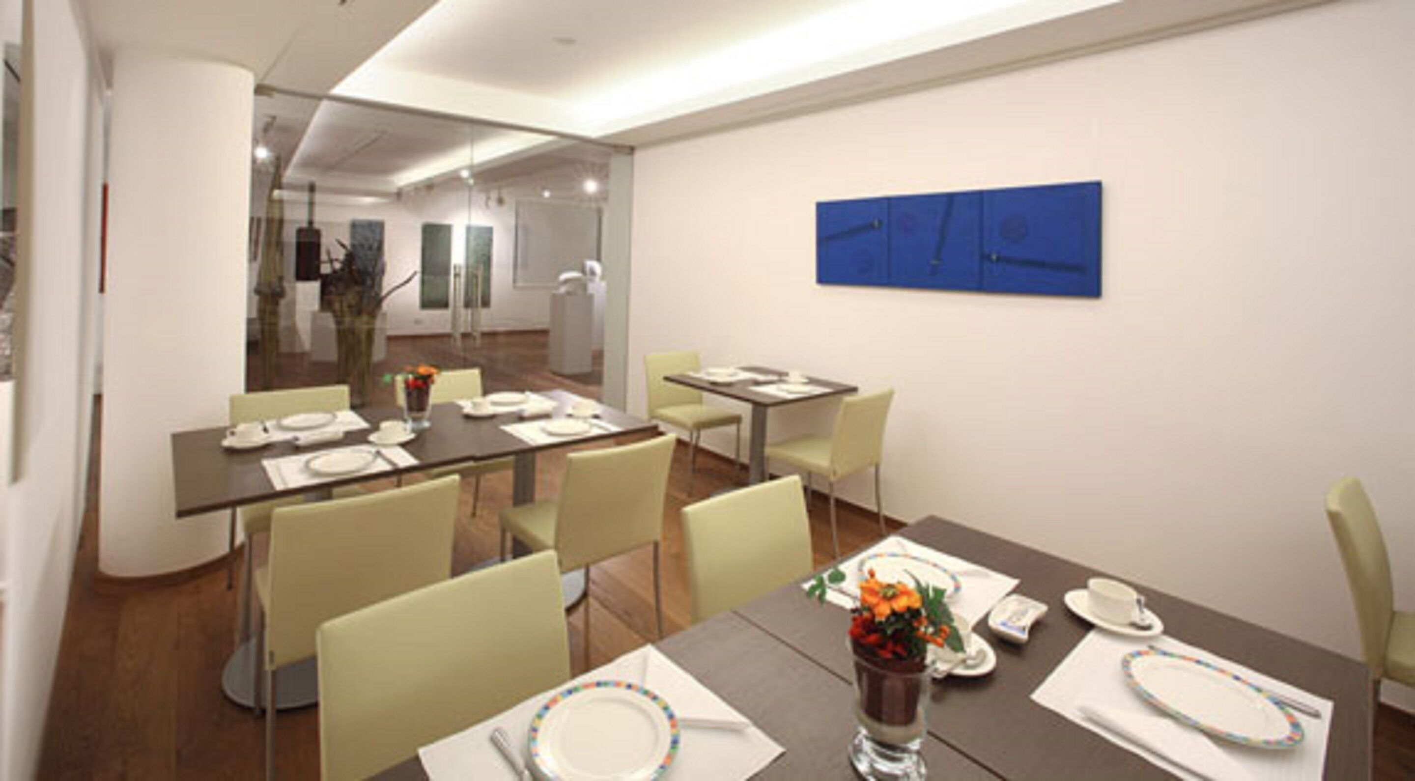 A modern conference room with glass walls, a blue abstract painting, and a dining area with tables set for four.