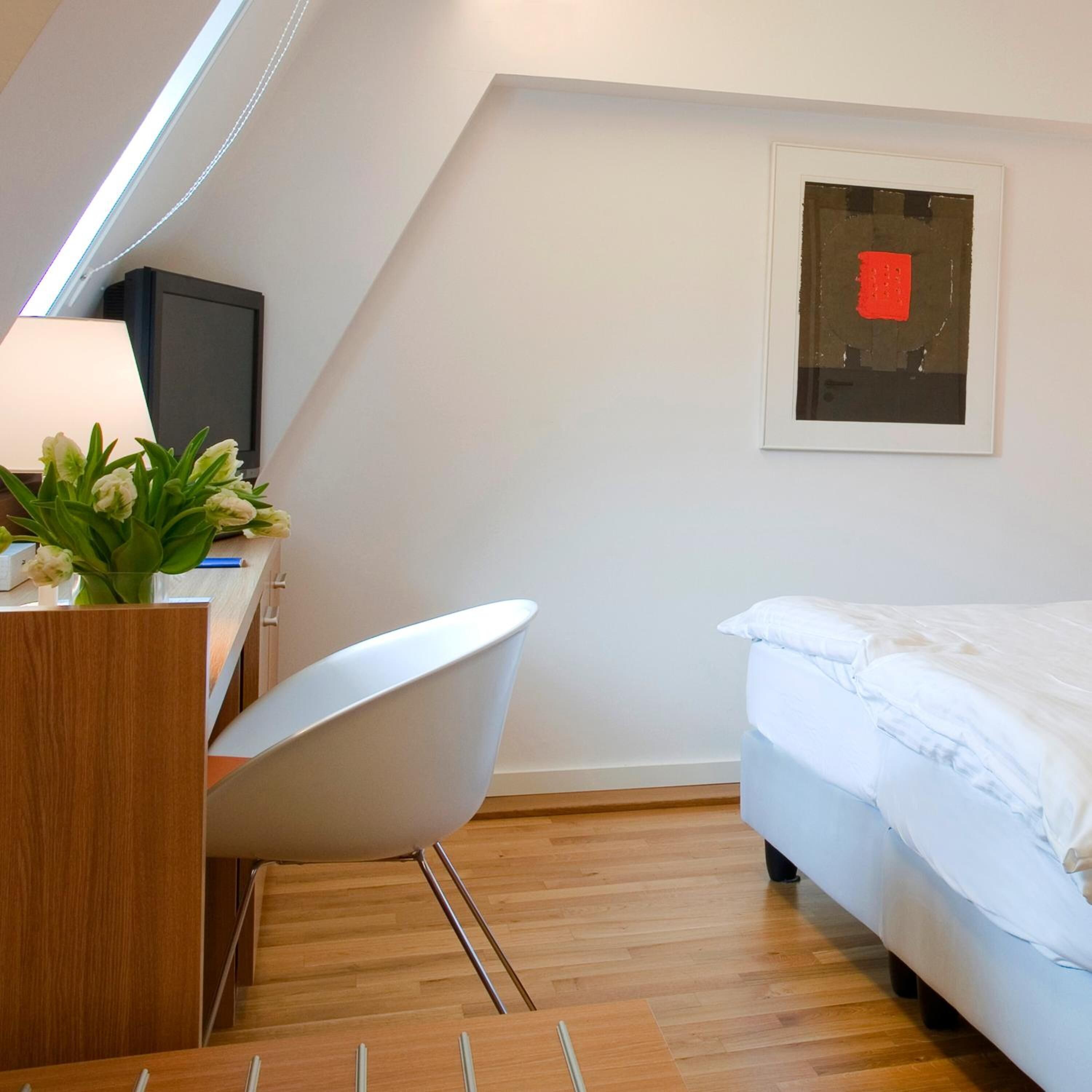 A modern bedroom with a sloped ceiling, a desk with a lamp, a chair, a bed, and a framed artwork on the wall.
