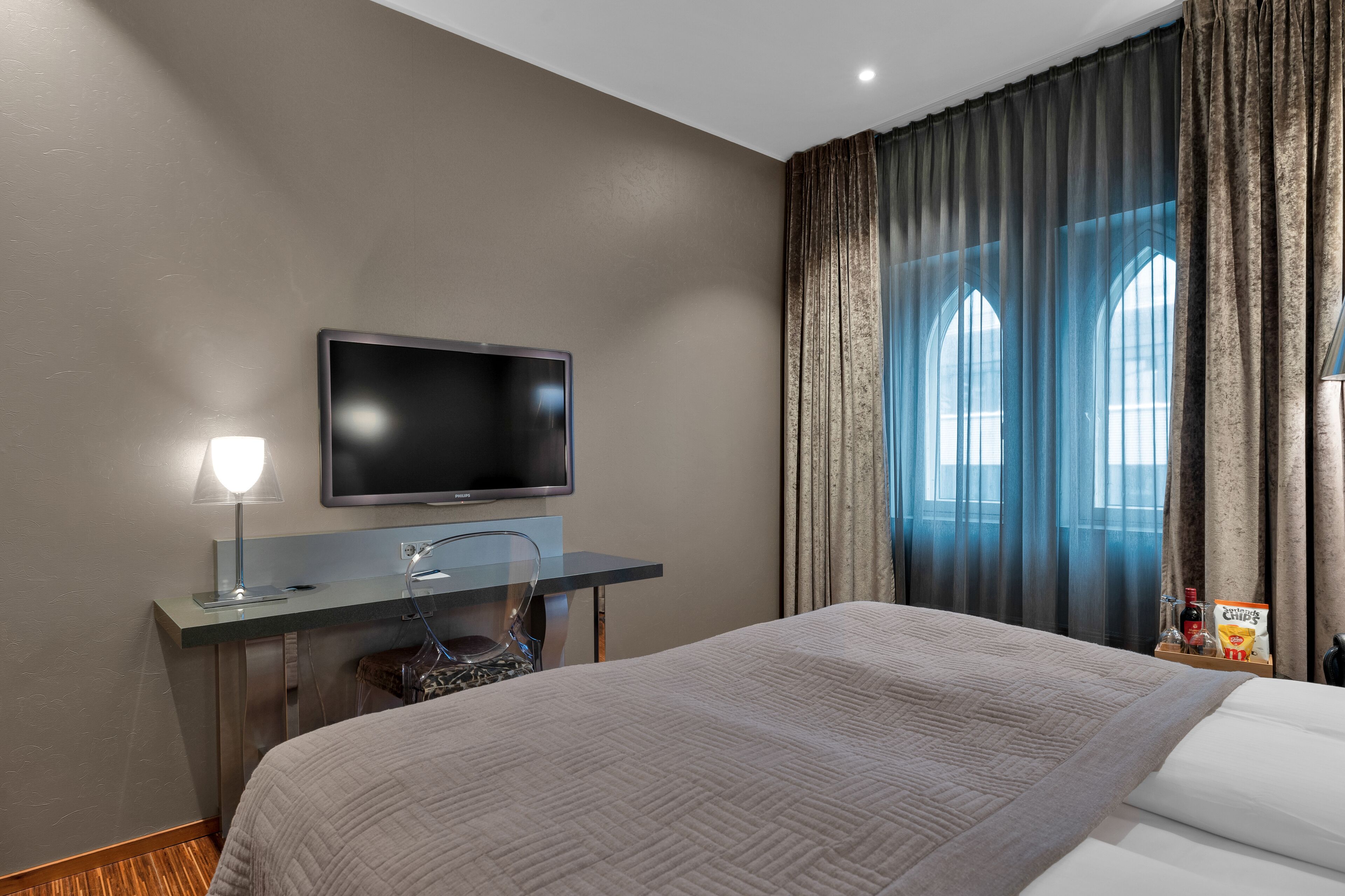 Una habitación de hotel con cama, escritorio con computadora, televisor y ventana con cortinas.