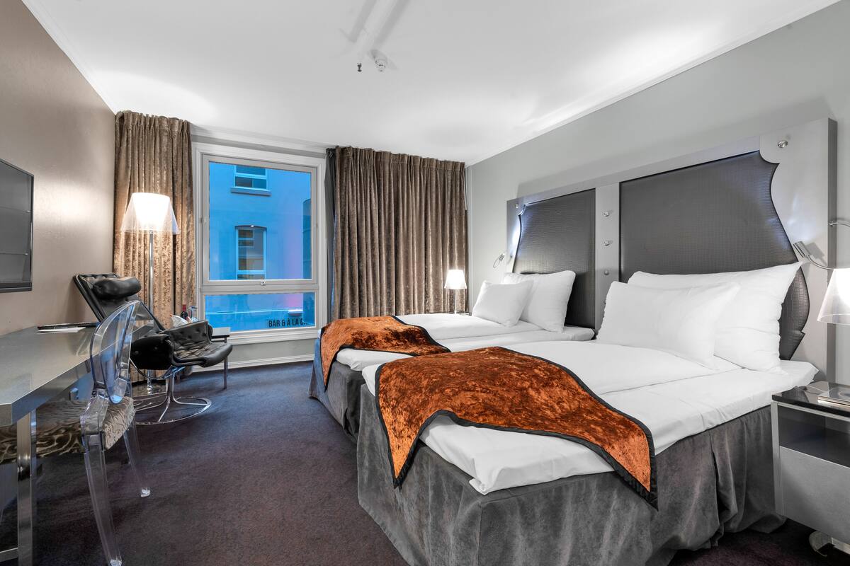 Ein Hotelzimmer mit zwei Betten, einem Schreibtisch, einem Stuhl und einem Fenster mit Blick auf ein Gebäude.