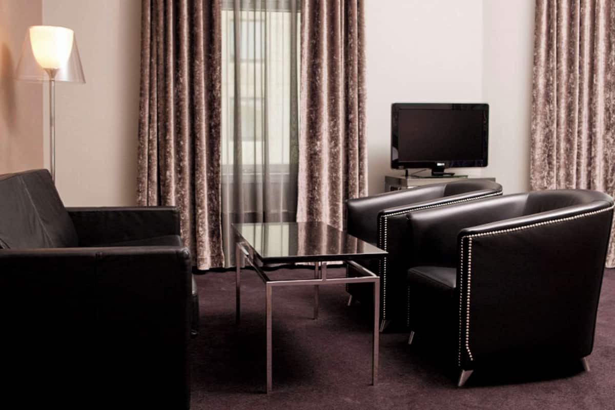 Ein modernes Hotelzimmer mit einer schwarzen Ledersofa, einem Glastisch, einem Flachbildfernseher und einem großen Fenster mit Vorhängen.