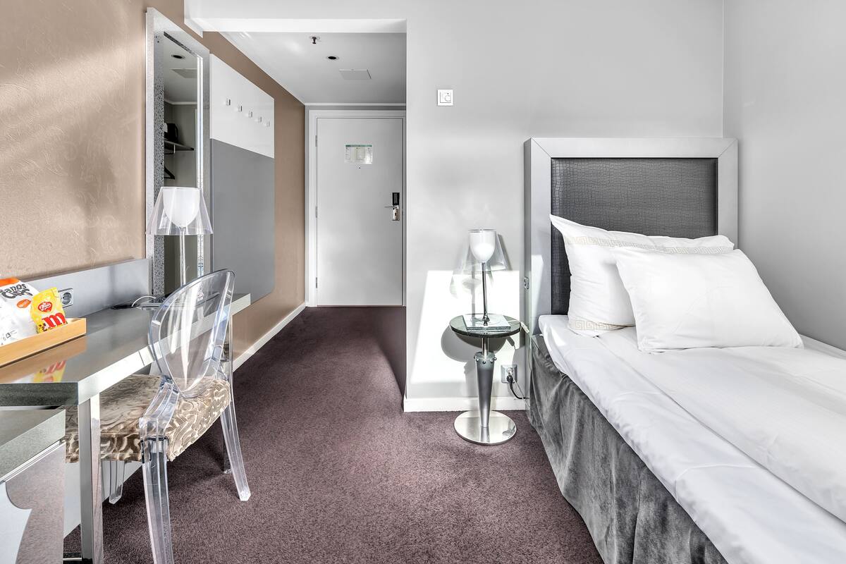 Ein Hotelzimmer mit einem Bett, einem Schreibtisch mit Stuhl, einem Spiegel, einem kleinen Tisch mit Lampe und einer Tür zu einem Badezimmer.