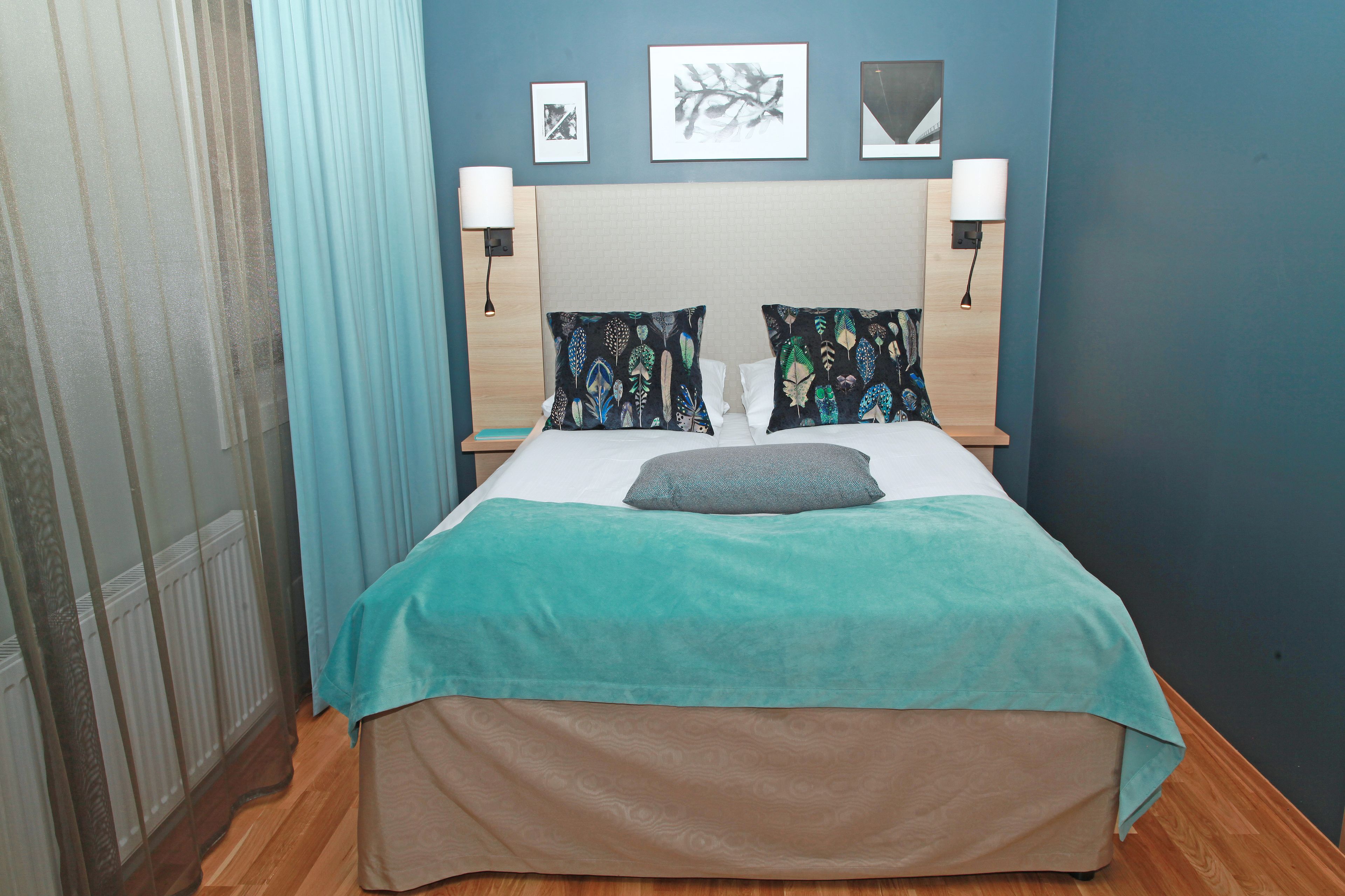 Una cama bien hecha con una colcha verde azulada y beige, dos almohadas decorativas y dos lámparas fijadas a la pared.