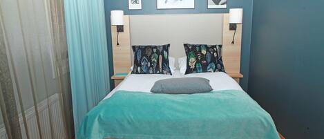 Una cama bien hecha con una colcha verde azulada y beige, dos almohadas decorativas y dos lámparas fijadas a la pared.