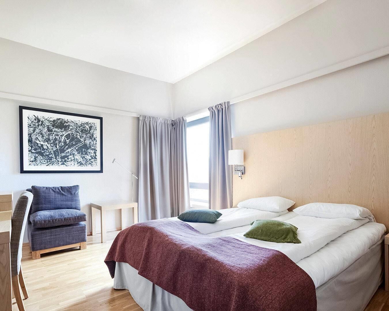 Et hotelværelse med seng, stol, skrivebord, et vindue med gardiner og en væglampe.