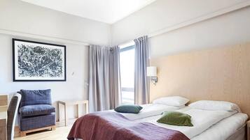Et hotelværelse med seng, stol, skrivebord, et vindue med gardiner og en væglampe.