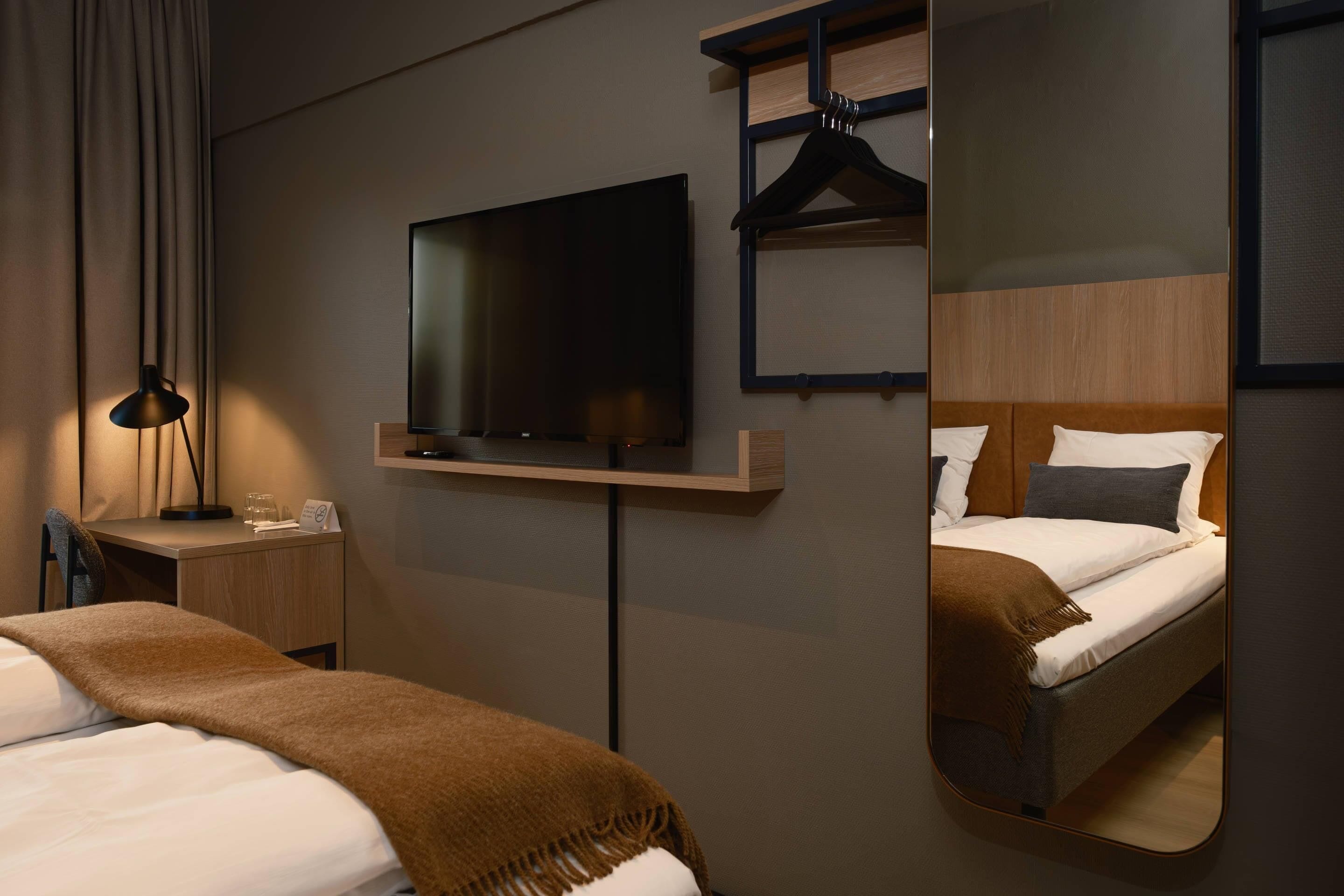 A hotel room with a bed, a TV mounted on the wall, a bedside table with a lamp, and a mirror reflecting the bed.