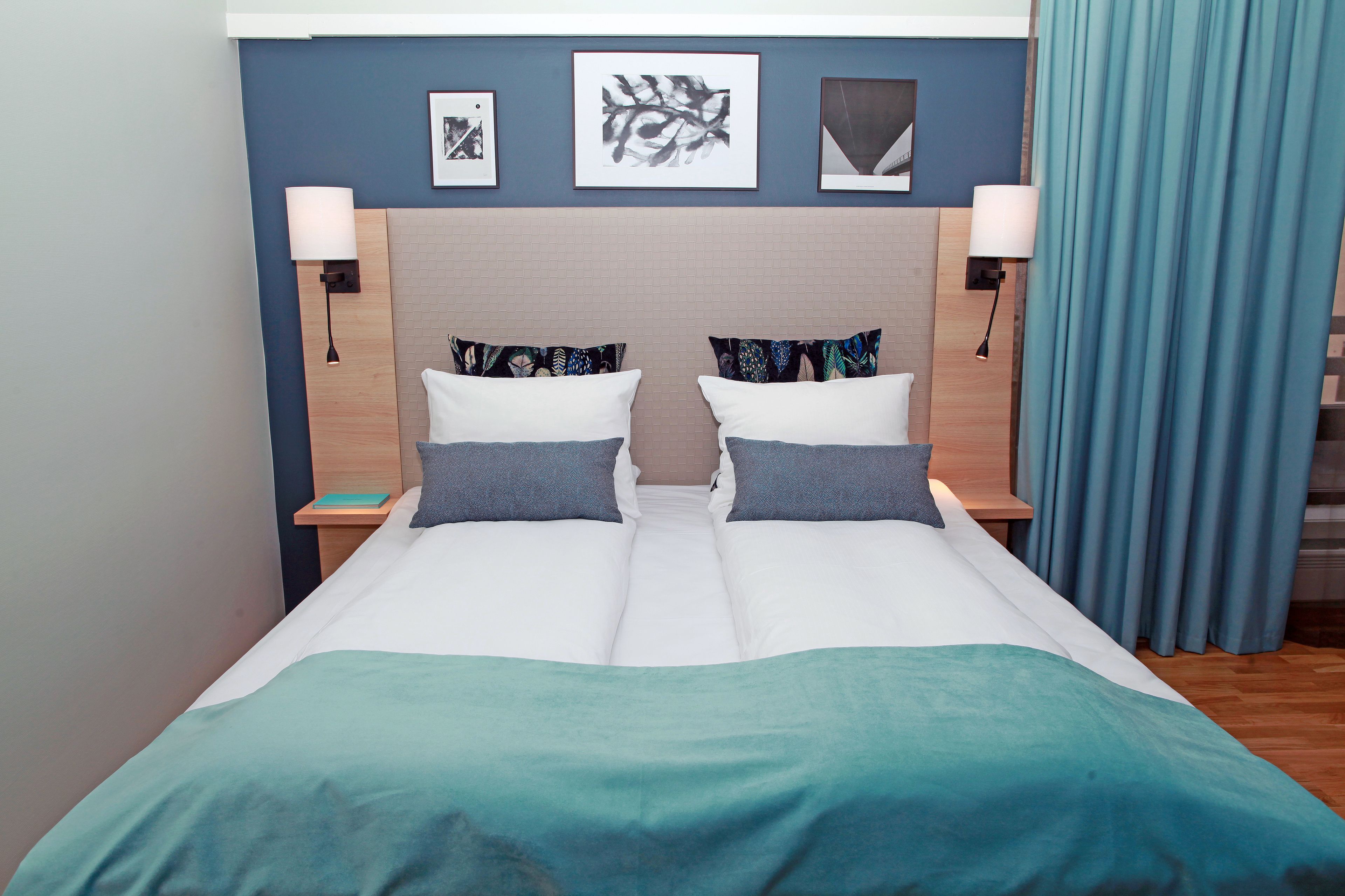 A double bed with a teal bedspread, two pillows, and two bedside tables with lamps. There are three framed pictures on the wall above the bed.