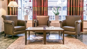 A hotel lobby with leather armchairs, a glass-top coffee table, and a view through large windows with red curtains. - Quality Hotel Grand Steinkjer (Steinkjer)