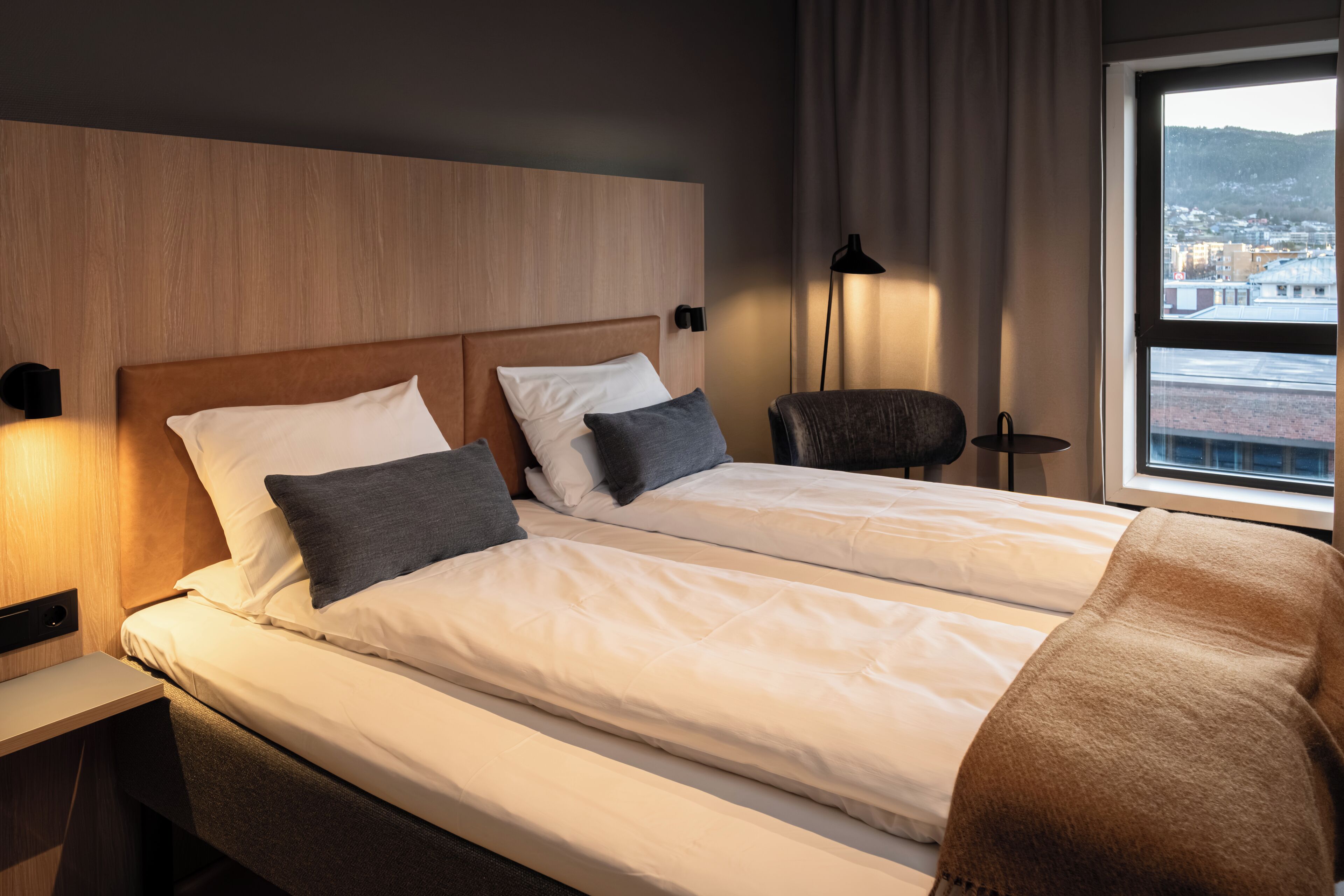 Une chambre d’hôtel avec deux lits, une tête de lit en bois, une chaise, une lampe et une fenêtre donnant sur la ville.