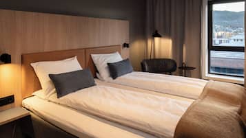 Ein Hotelzimmer mit zwei Betten, einem Holz-Kopfteil, einem Sessel, einer Lampe und einem Fenster mit Blick auf die Stadt.