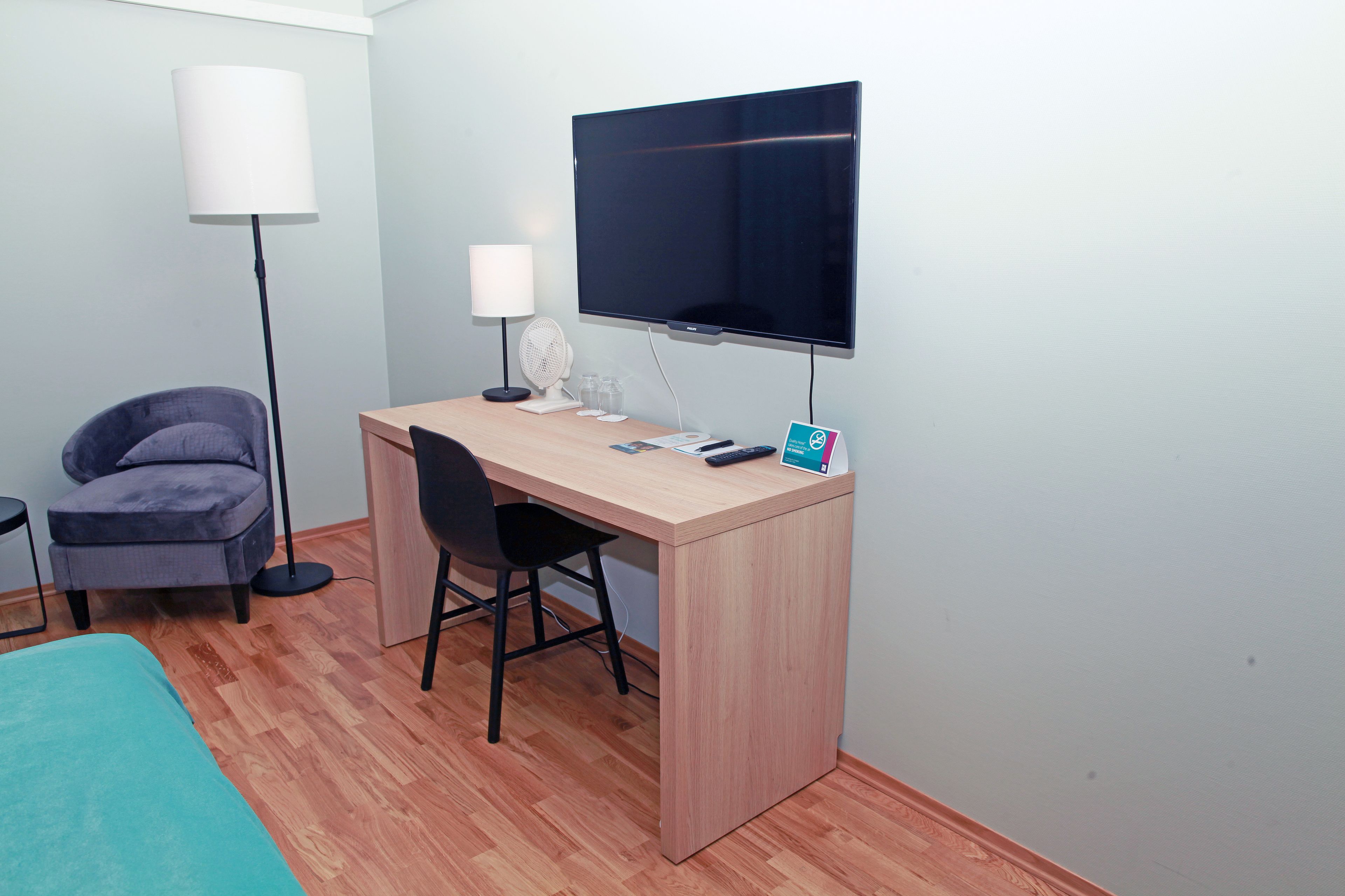 A small, modern room with a wooden desk, a chair, a flat-screen TV, a lamp, and a small table with a glass.