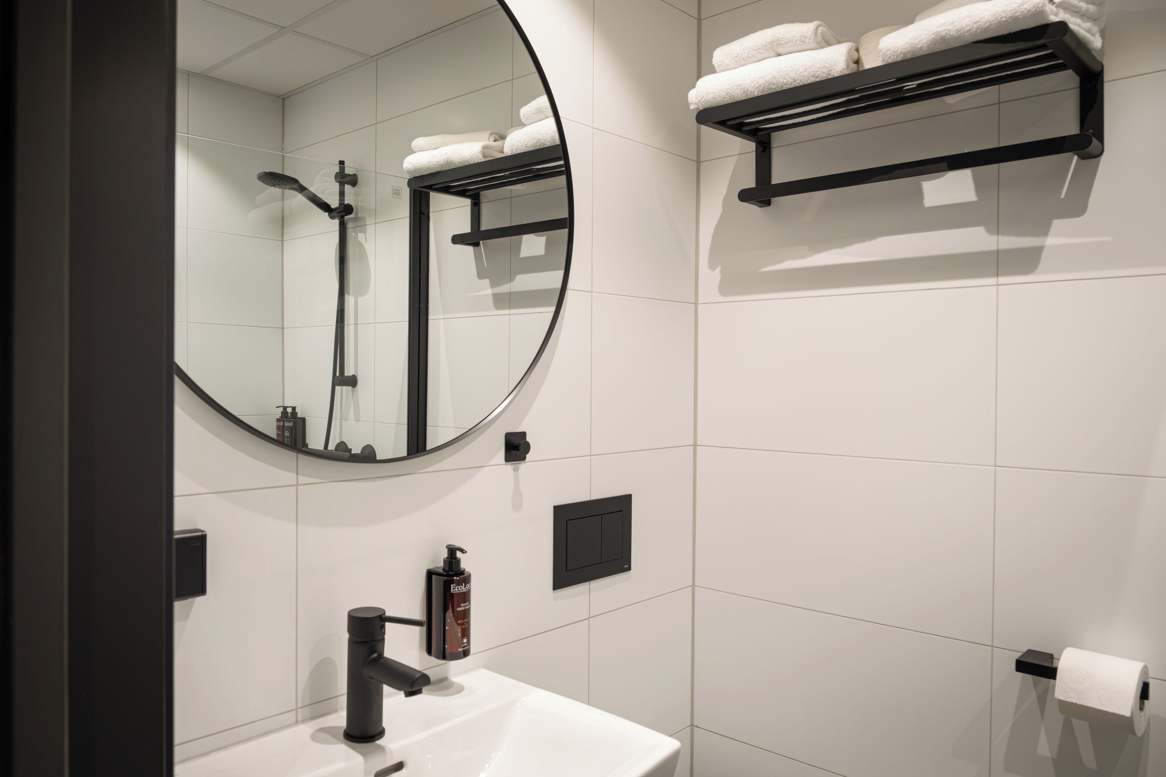 A modern bathroom with a round mirror, a black towel rack, a white sink, and a toilet paper holder.