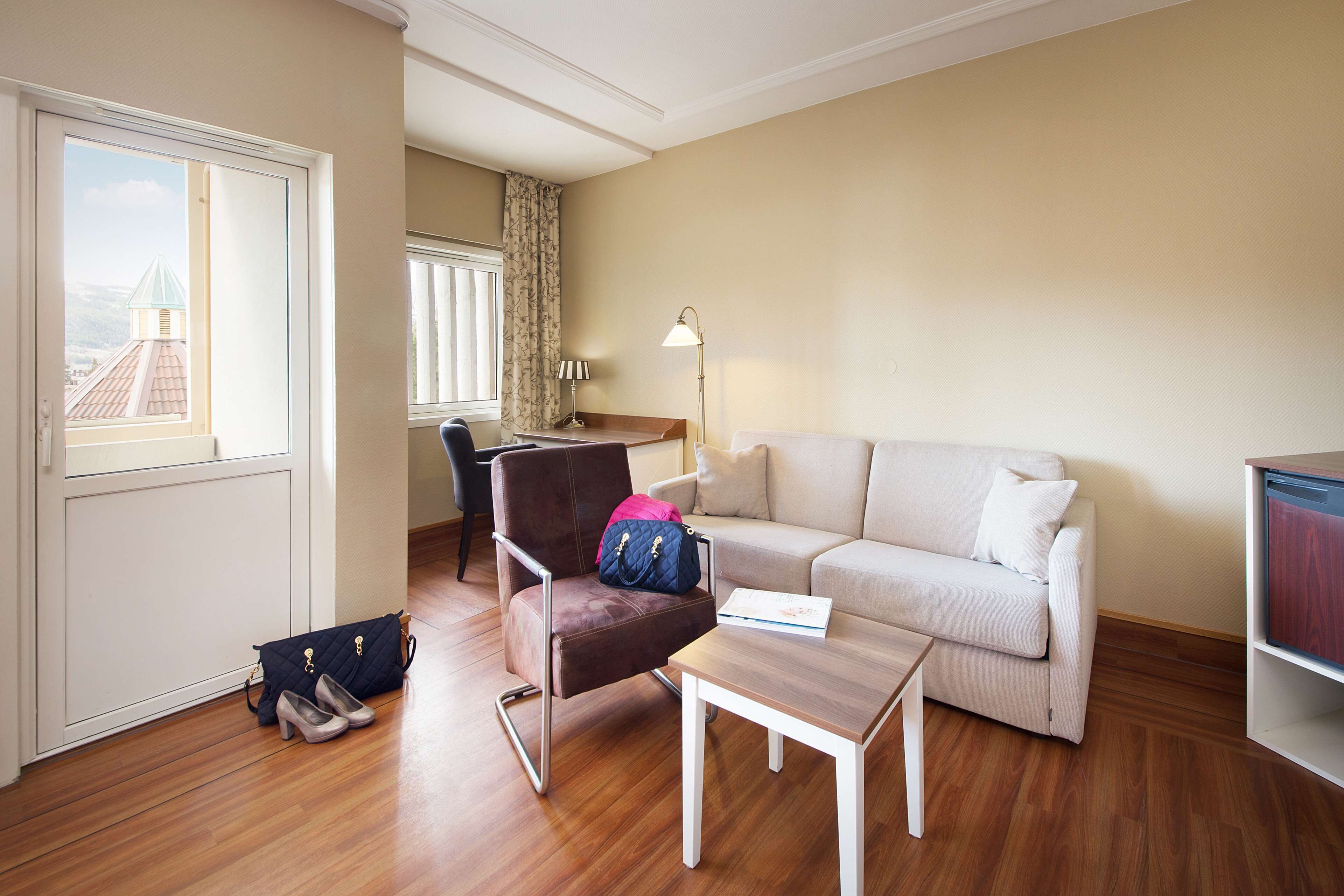 A living room with a wooden floor, a sofa, a chair, a small table, a lamp, and a window with a view of a building.