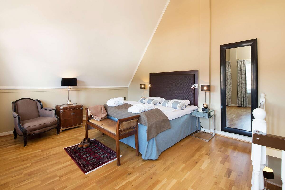 A bedroom with a bed, a chair, a small table, a lamp, a mirror, and a window.