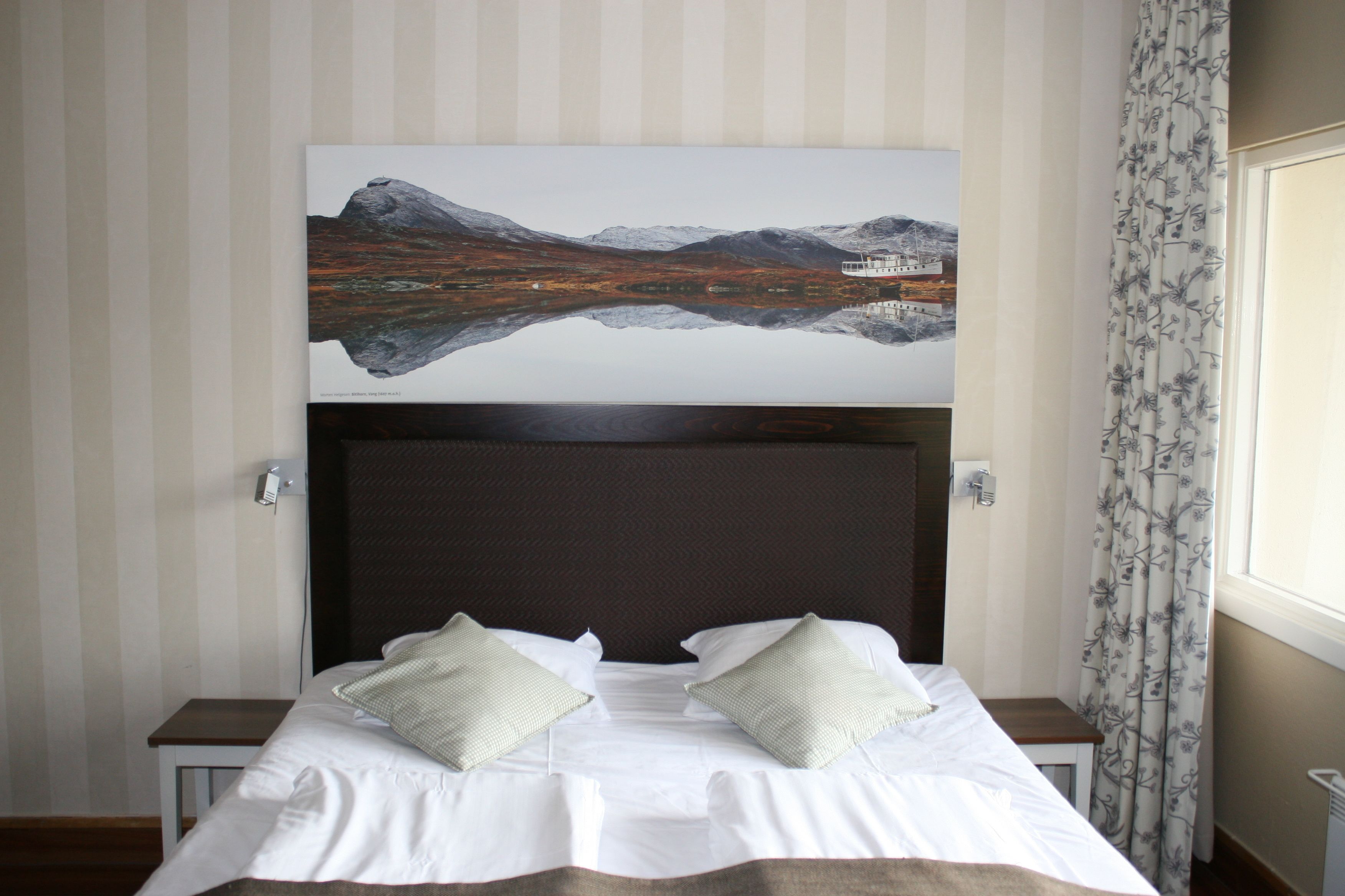 A neatly made bed with white linens and two pillows, a wooden headboard, and a painting of a mountainous landscape above the bed.