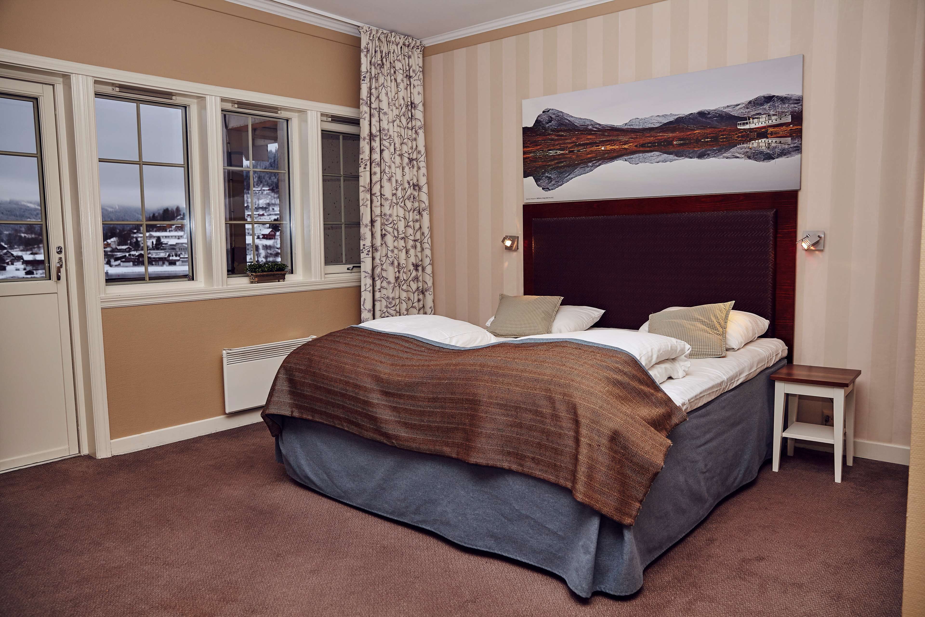 A bedroom with a large bed, a view of a snowy landscape through the window, and a painting of a mountain landscape on the wall.