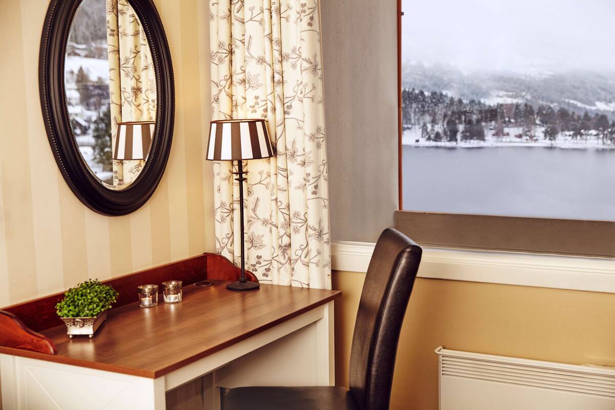 A cozy room with a wooden desk, a chair, a lamp, a mirror, and a view of a snowy landscape through a window.