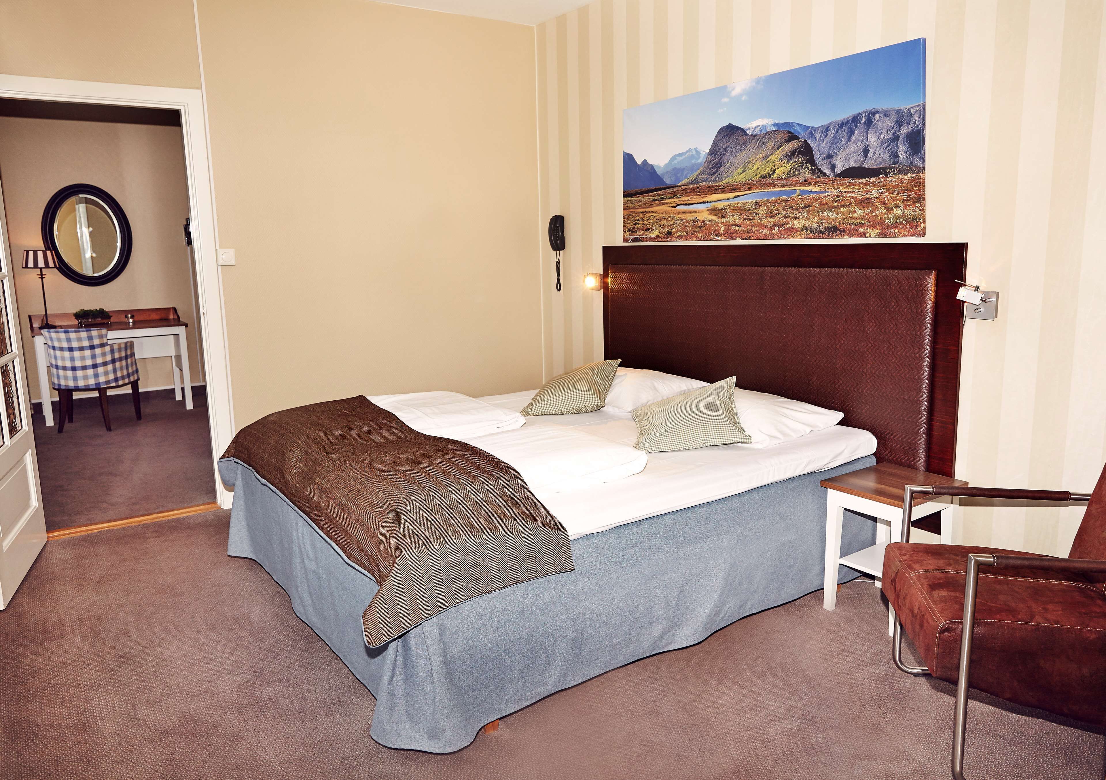 Ett hotellrum med en stor säng, en sänggavel i trä, ett litet sidobord, en stol och en tavla med ett bergigt landskap på väggen.