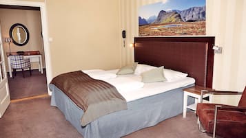 Een hotelkamer met een groot bed, een houten hoofdbord, een klein nachtkastje, een stoel en een schilderij van een berglandschap aan de muur.