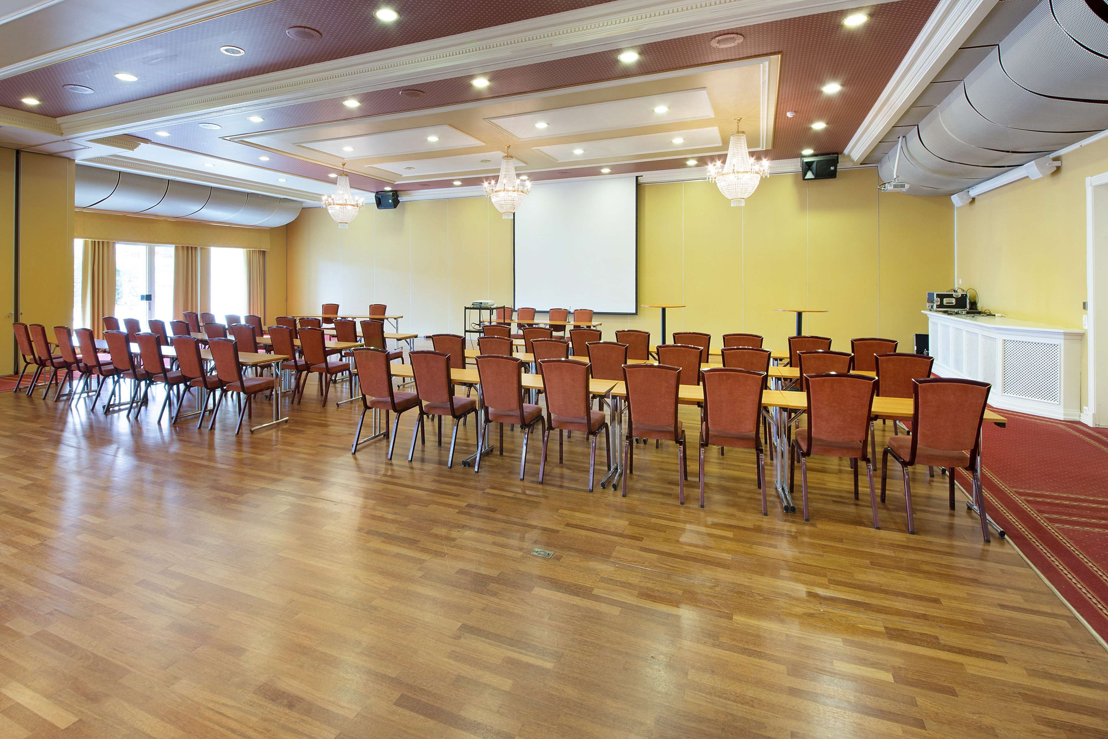 Uma sala de conferências com fileiras de cadeiras e mesas, uma tela de projetor e piso coberto por carpete.