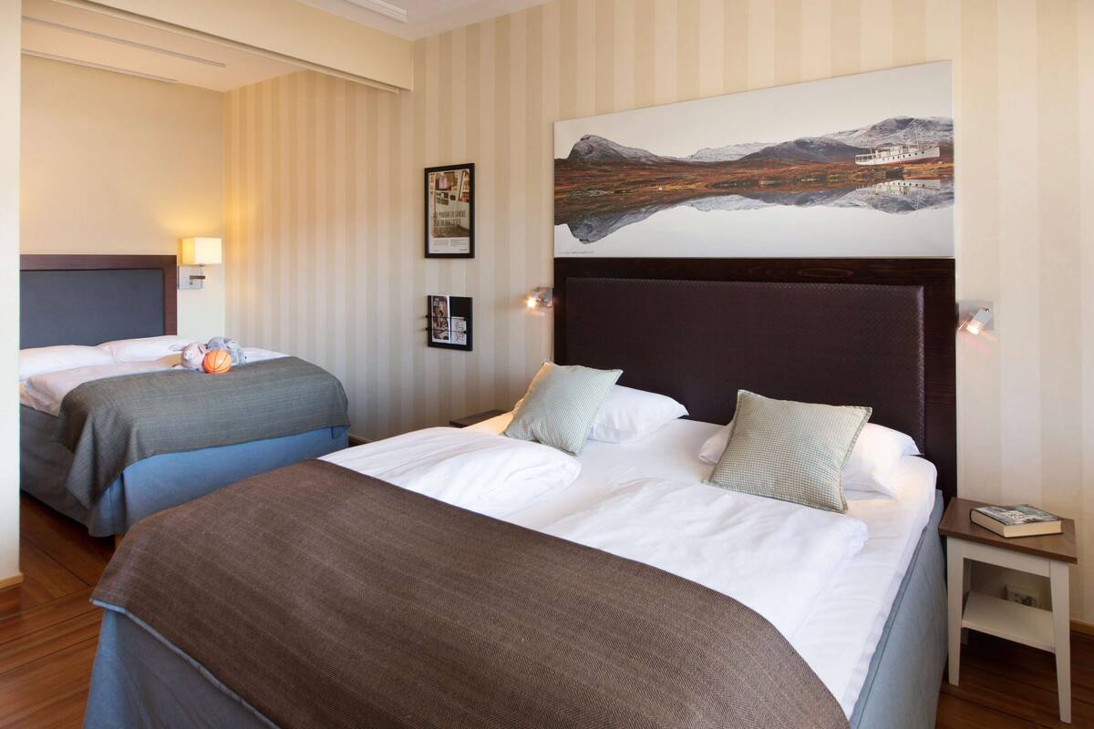 A hotel room with two beds, a large painting of a mountainous landscape, and a small bedside table with a book.