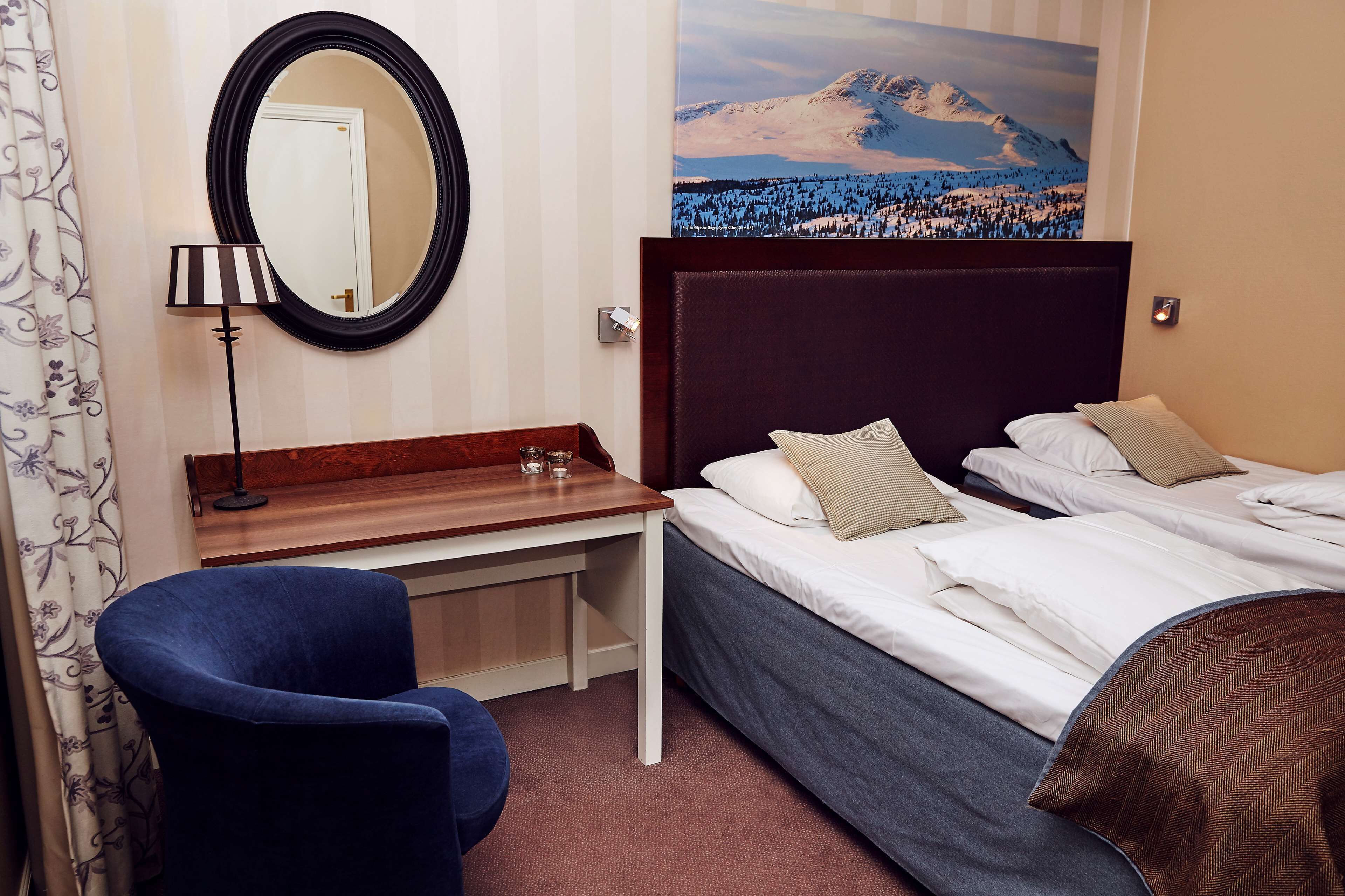 Ein Hotelzimmer mit Bett, Schreibtisch, Stuhl, Spiegel und einem Gemälde einer verschneiten Berglandschaft.