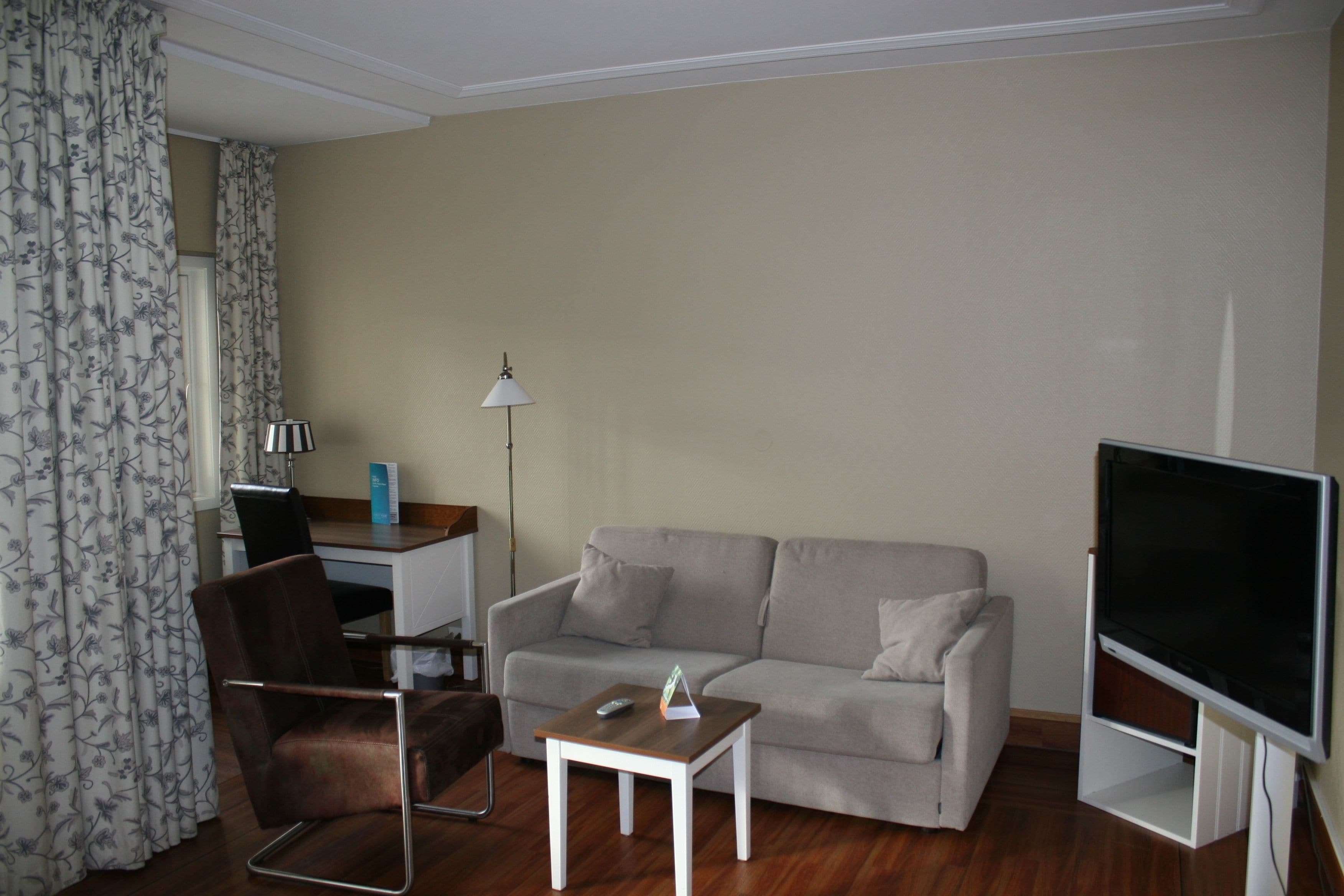 Een woonkamer met een bank, fauteuil, salontafel, televisie en een bureau met een lamp.
