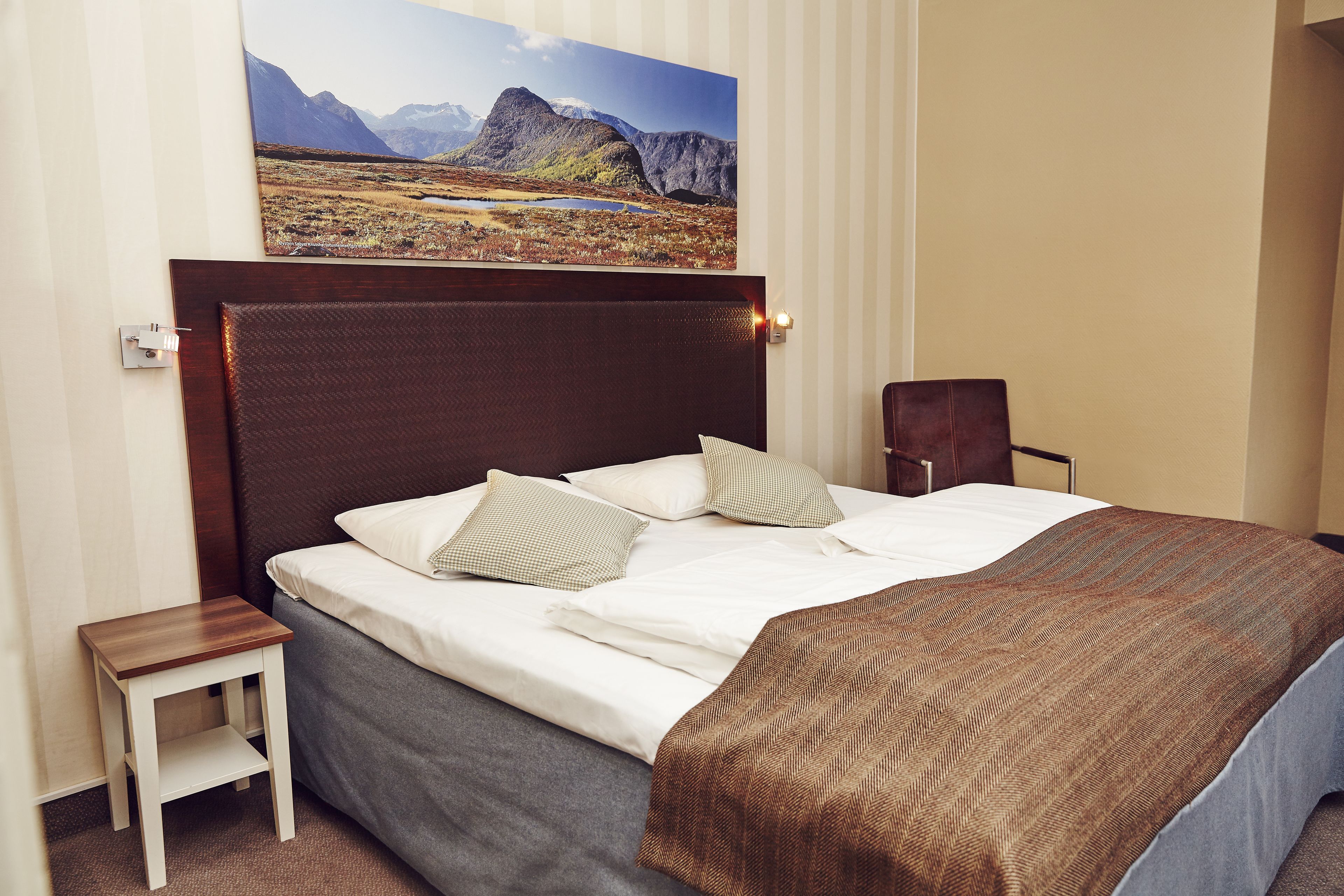 Ein ordentlich bezogenes Bett mit brauner Decke und Kissen, ein kleiner Nachttisch aus Holz und ein Gemälde einer Berglandschaft darüber.