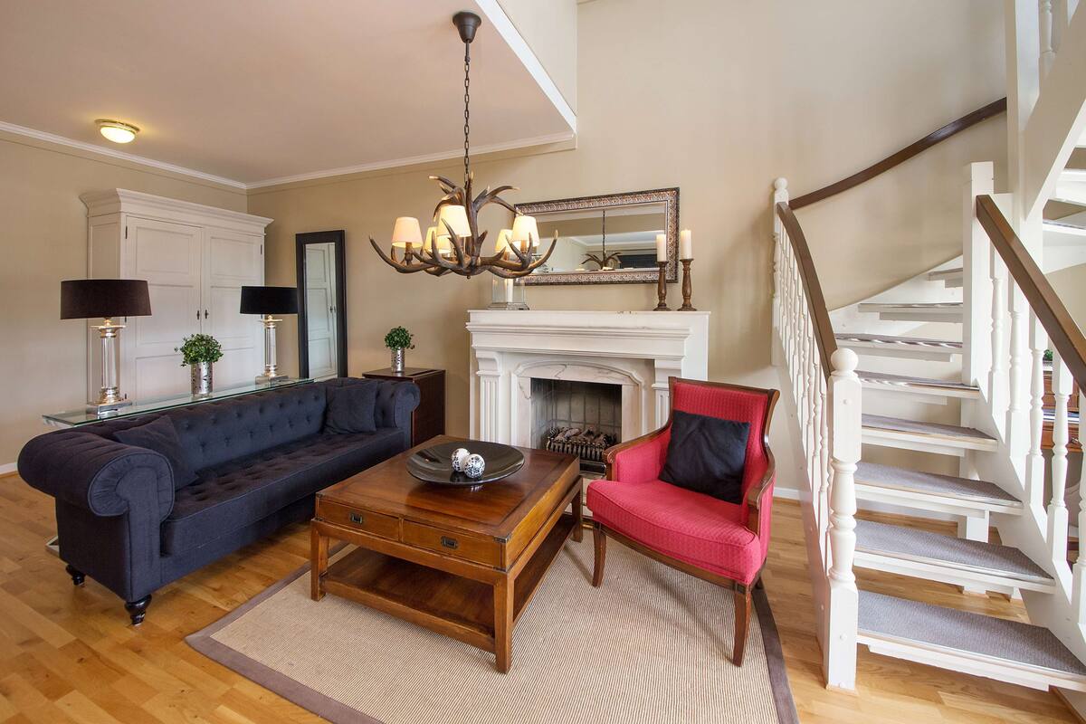 A living room with a dark blue sofa, a wooden coffee table, a red armchair, a fireplace, and a white staircase.