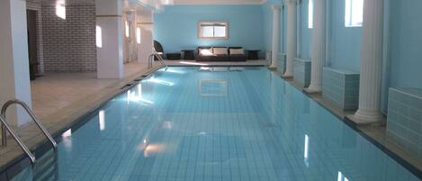 A long, rectangular indoor swimming pool with clear blue water, surrounded by white columns and a blue wall.