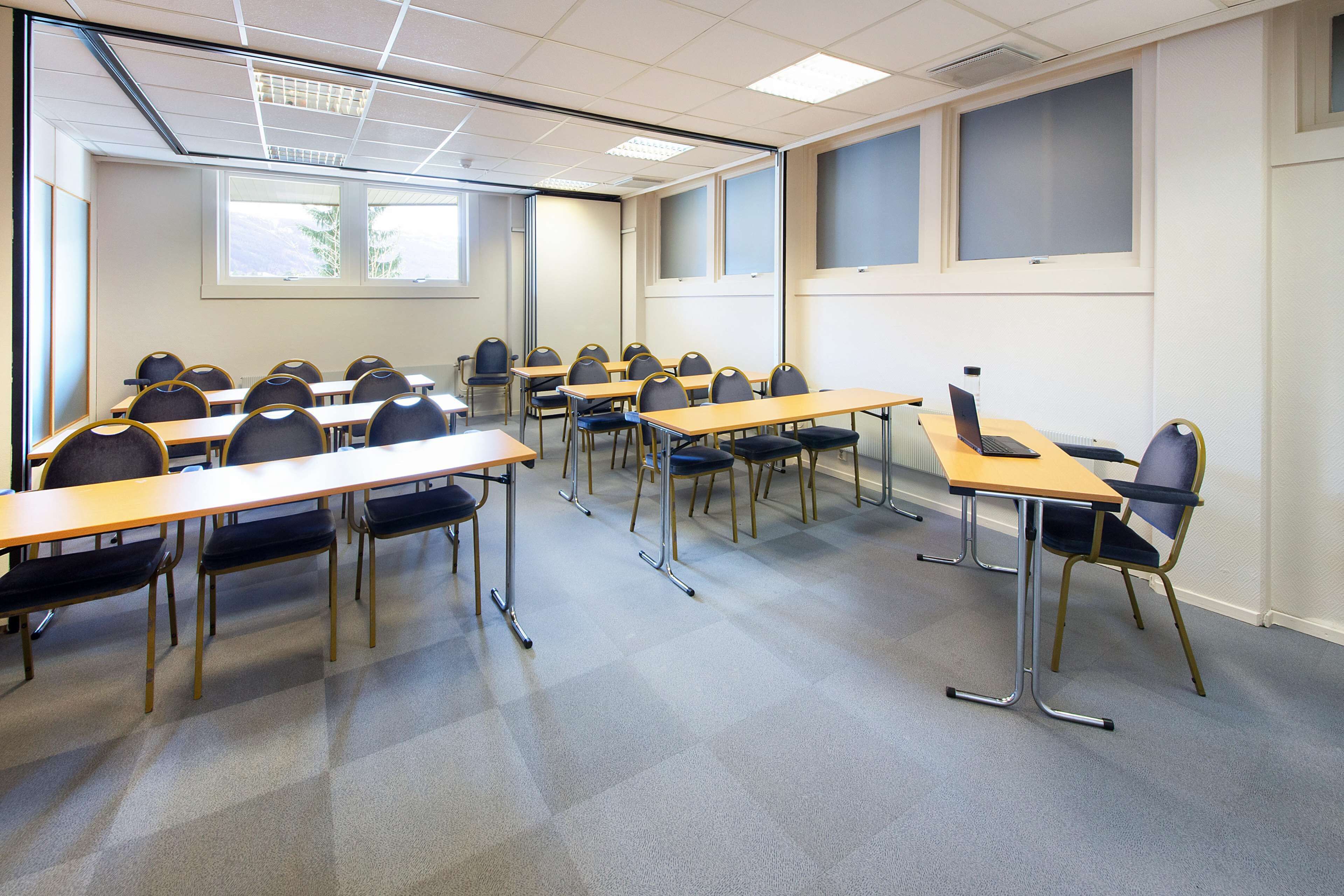 Een vergaderruimte met meerdere tafels en stoelen, grote ramen en een projectiescherm.