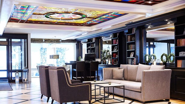 A spacious lobby with a large sofa, armchairs, and a coffee table. There are bookshelves filled with books and decorative items. A stained glass ceiling adds a unique touch to the space. - Scandic Valdres (Nord-Aurdal)