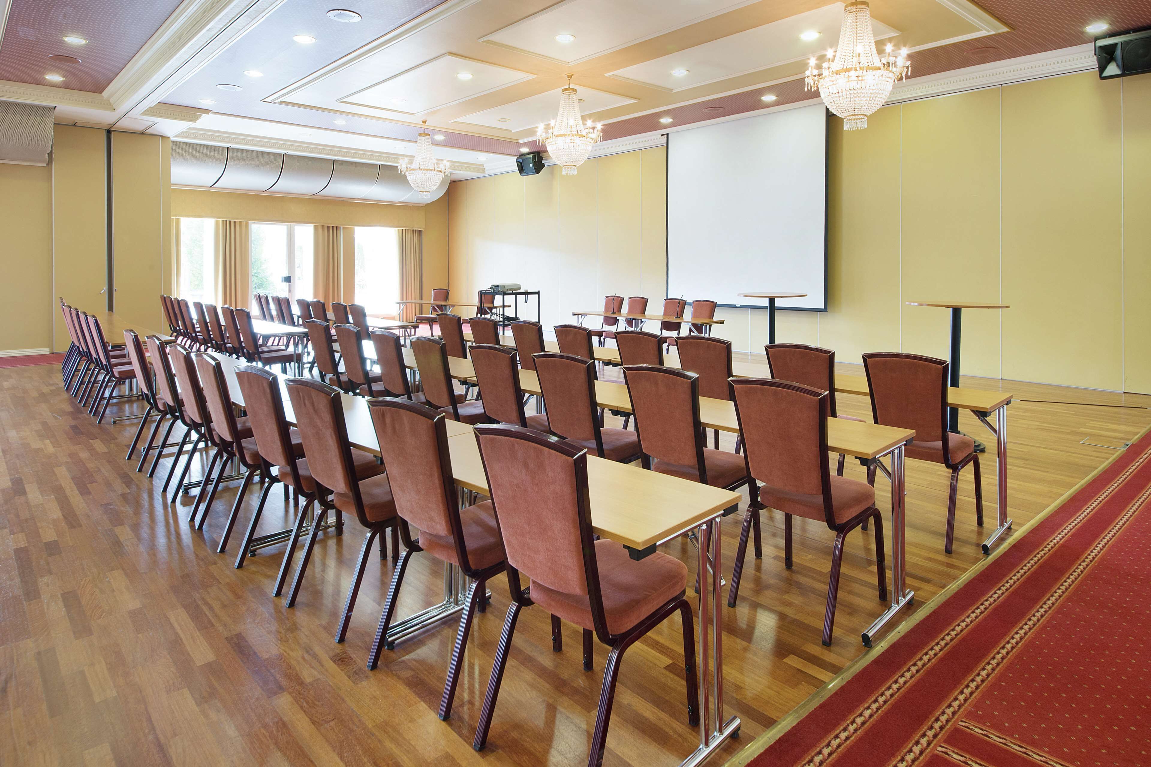 Uma sala de conferências com fileiras de cadeiras e mesas, uma tela de projetor e lustres.