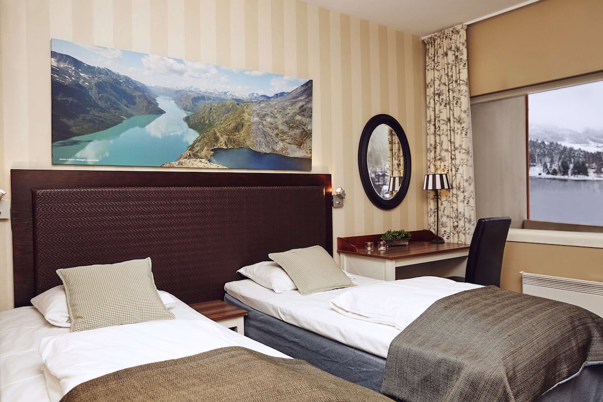 A hotel room with two beds, a large landscape painting, a mirror, and a view of a lake through the window.