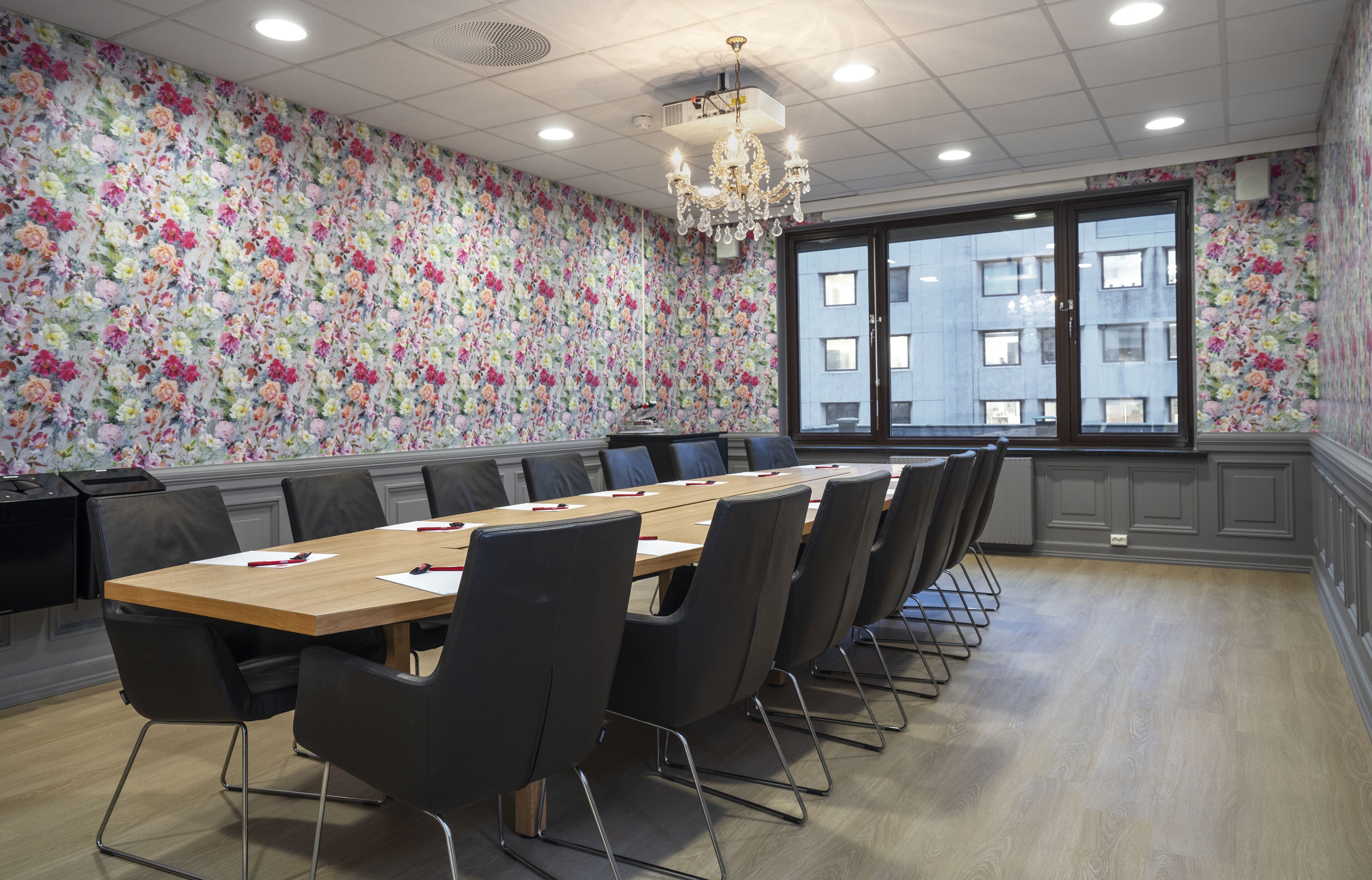 A conference room with a long table, black chairs, floral wallpaper, a chandelier, and a large window with a view of buildings.