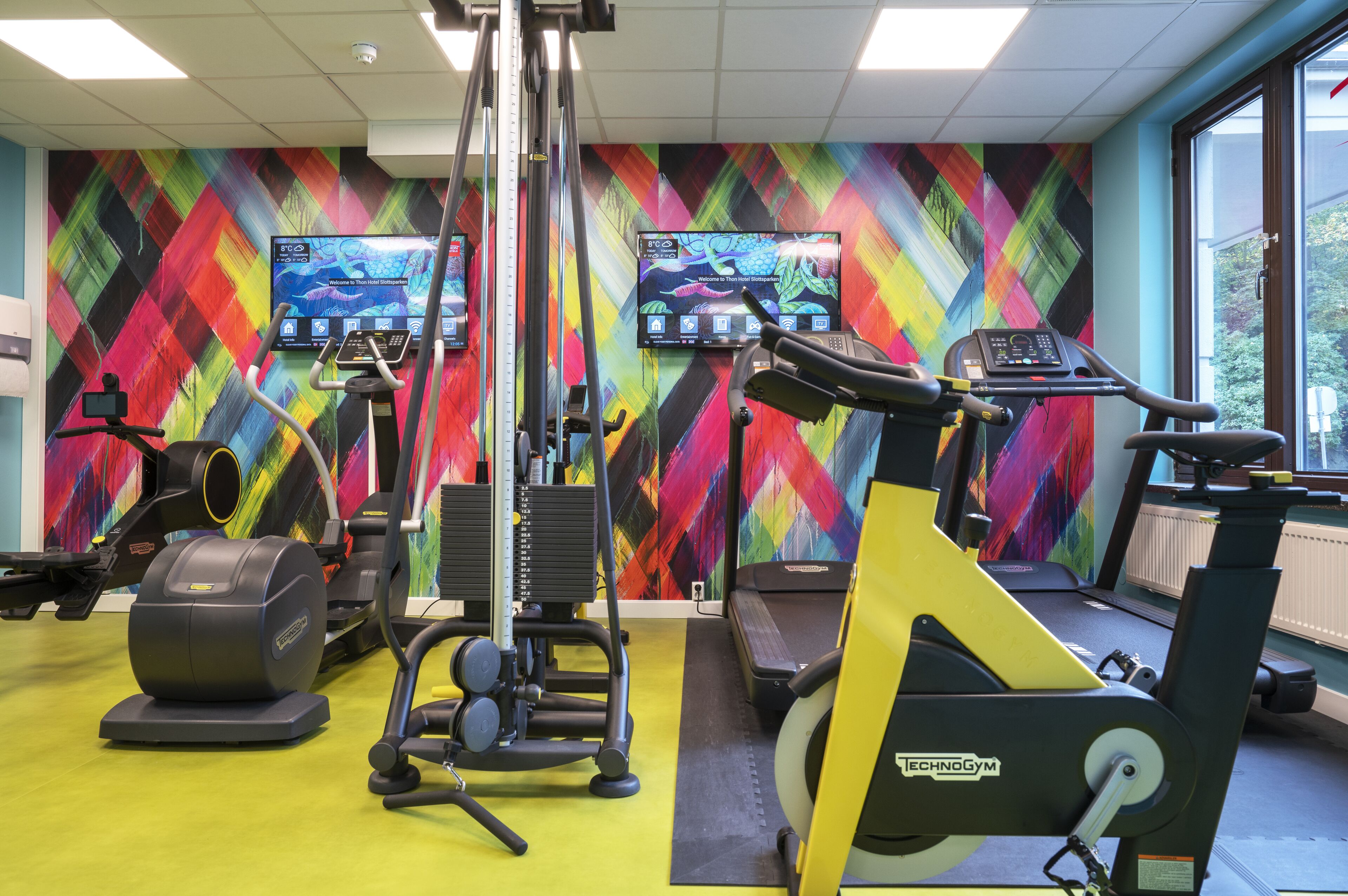 Un gimnasio con equipo de ejercicio, un mural de pared colorido y un ventanal que proporciona luz natural.