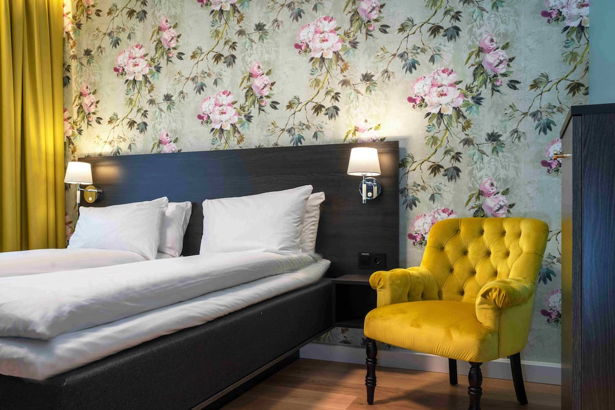 Una habitación de hotel con una cama, dos lámparas de noche, un sillón amarillo y papel tapiz con motivos florales.