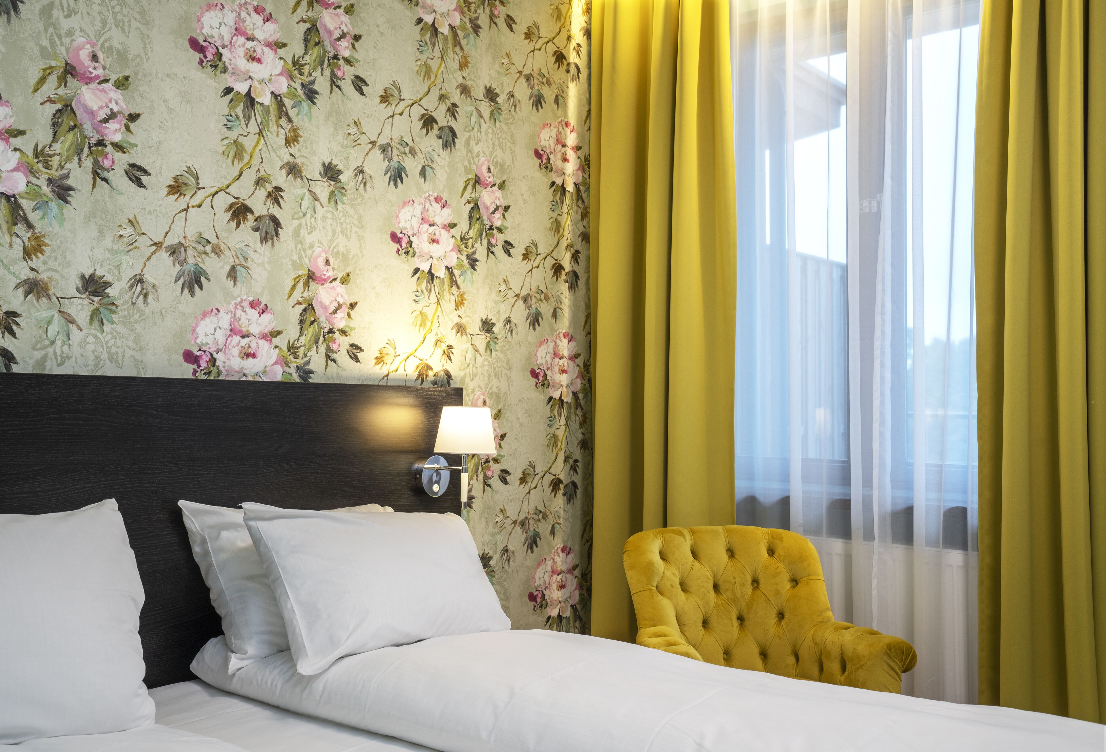Habitación de hotel con cama, mesita de noche con lámpara, papel tapiz floral y cortinas amarillas.