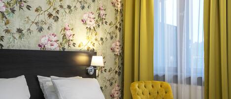 Habitación de hotel con cama, mesita de noche con lámpara, papel tapiz floral y cortinas amarillas.