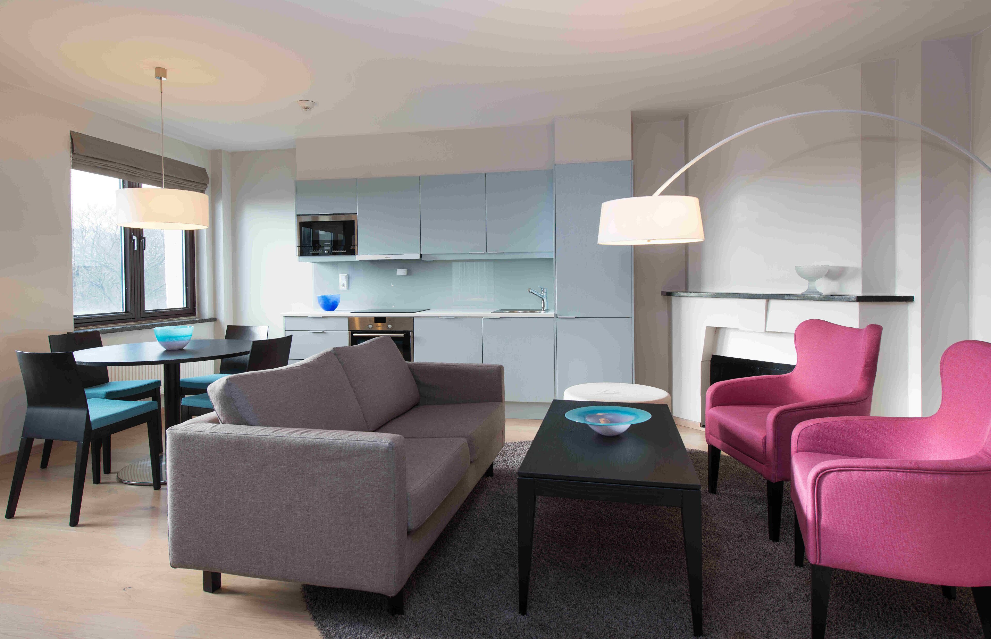 A modern living room with a grey sofa, two pink armchairs, a coffee table, and a dining area with a table and chairs.
