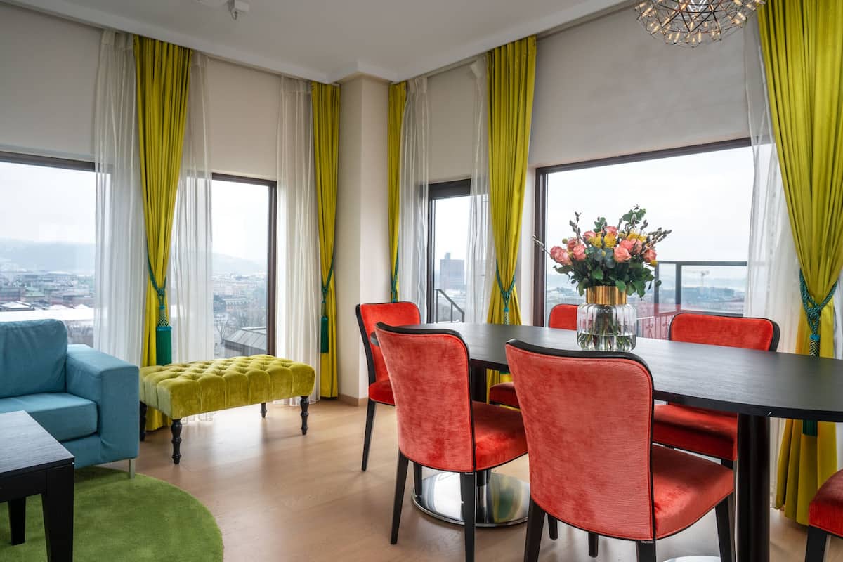 Una sala de estar moderna con un sofá, una mesa de comedor con sillas y vistas a un paisaje urbano a través de grandes ventanales.