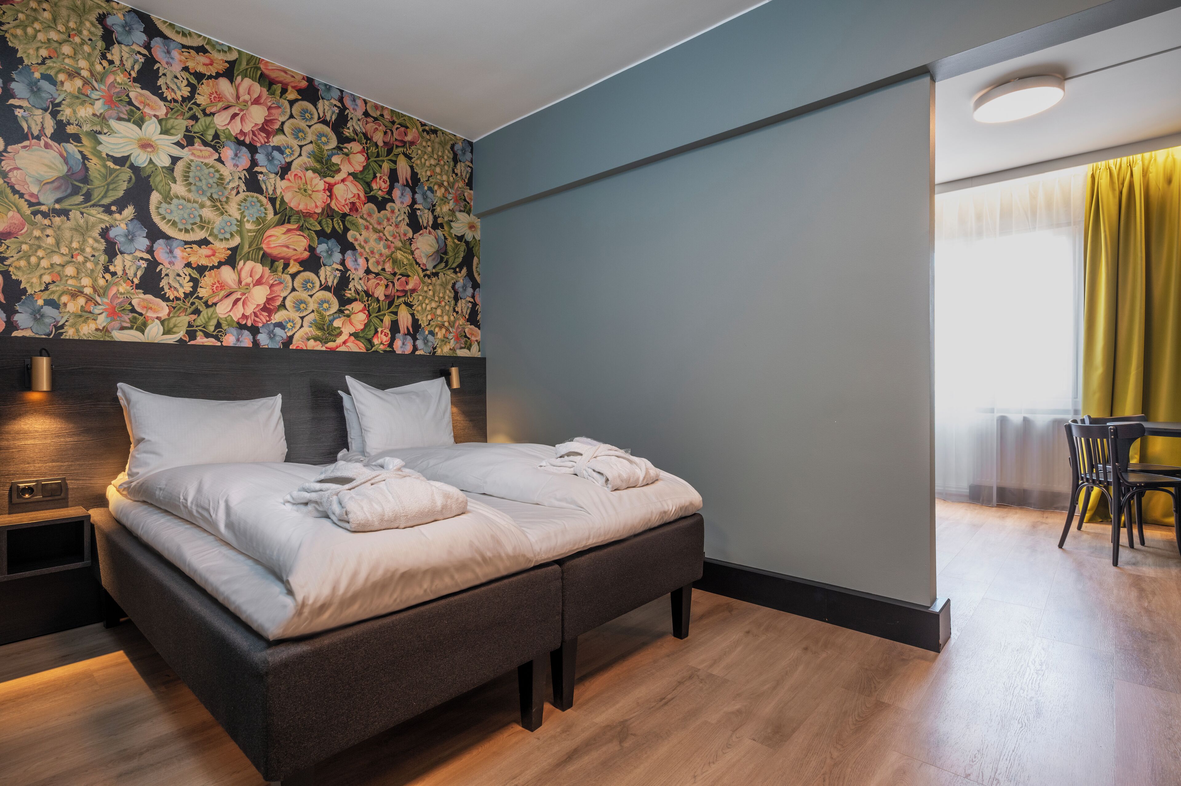 Une chambre d’hôtel moderne avec un grand lit, un papier peint à motifs floraux, une table de chevet avec une lampe et une vue sur un espace repas.